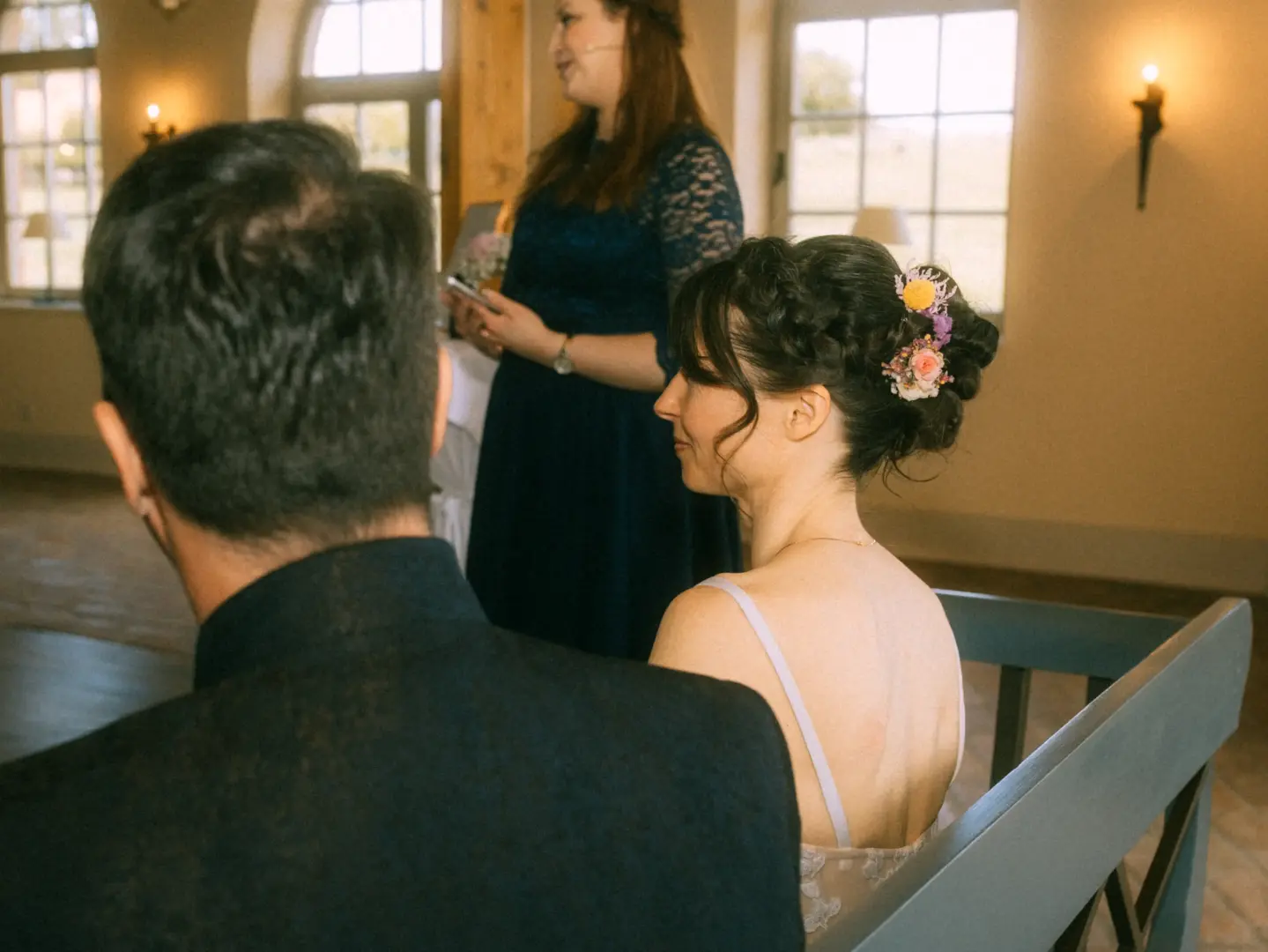 Eine Braut mit kunstvoller Hochsteckfrisur und Blumenschmuck sitzt neben ihrem Bräutigam während einer Trauung, während eine Rednerin im Hintergrund spricht. Stimmungsvolles Bild vom Hochzeitsfotograf in Schwerin und MV.
