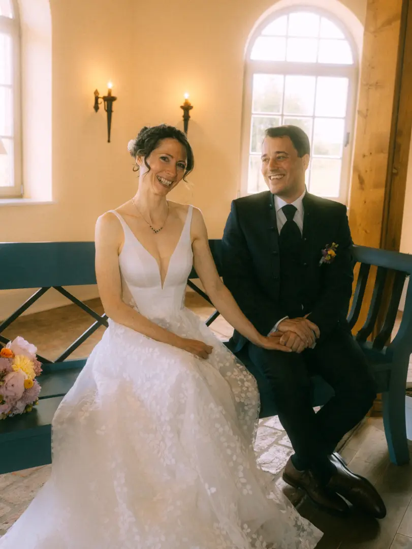 Ein glückliches Brautpaar sitzt Händchen haltend auf einer blauen Holzbank in einem hellen Raum. Die Braut trägt ein weißes Spitzenkleid, der Bräutigam einen eleganten schwarzen Anzug. Ein Lovestory Fotograf in Schwerin und MV hat diesen intimen Moment der Hochzeit festgehalten.