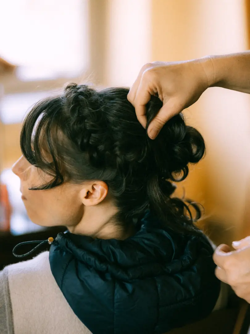 Eine Braut erhält von ihrem Hochzeitsfotograf in Schwerin und MV ein kunstvolles Brautstyling mit geflochtenen Details. Die Stylistin steckt ihr dunkles Haar liebevoll hoch, während die Braut in einem Profilblick entspannt in die Ferne schaut.