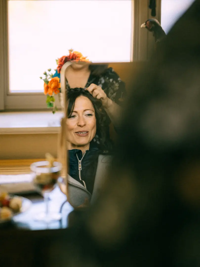 Eine Braut betrachtet sich lächelnd im Spiegel, während eine Stylistin ihr die Haare für den großen Tag frisiert. Im Hintergrund sind farbenfrohe Blumenarrangements zu sehen. Eine authentische Aufnahme vom Hochzeitsfotograf in Schwerin und MV.
