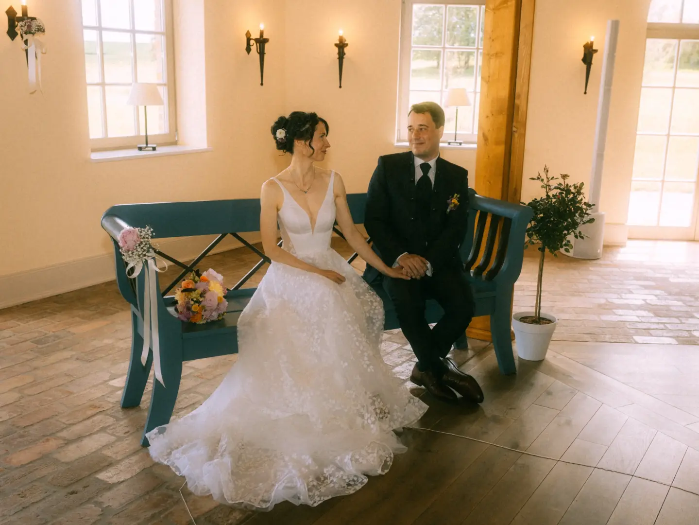 Ein Brautpaar sitzt sich liebevoll anblickend auf einer blauen Holzbank in einem hellen, rustikalen Raum. Die Braut trägt ein weißes Spitzenkleid, der Bräutigam einen dunklen Anzug. Ein Hochzeitsfotograf in Schwerin und MV hat diesen Moment der Verbundenheit stimmungsvoll festgehalten.