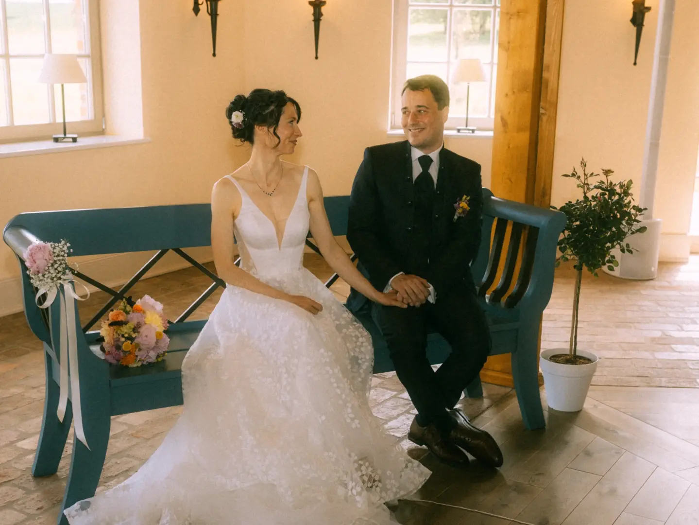 Ein frisch vermähltes Brautpaar sitzt sich liebevoll anblickend auf einer hellblauen Holzbank. Die Braut trägt ein elegantes weißes Spitzenkleid, der Bräutigam einen dunklen Anzug. Ein Hochzeitsfotograf in Schwerin und MV hat diesen romantischen Moment in einer lichtdurchfluteten Location festgehalten.