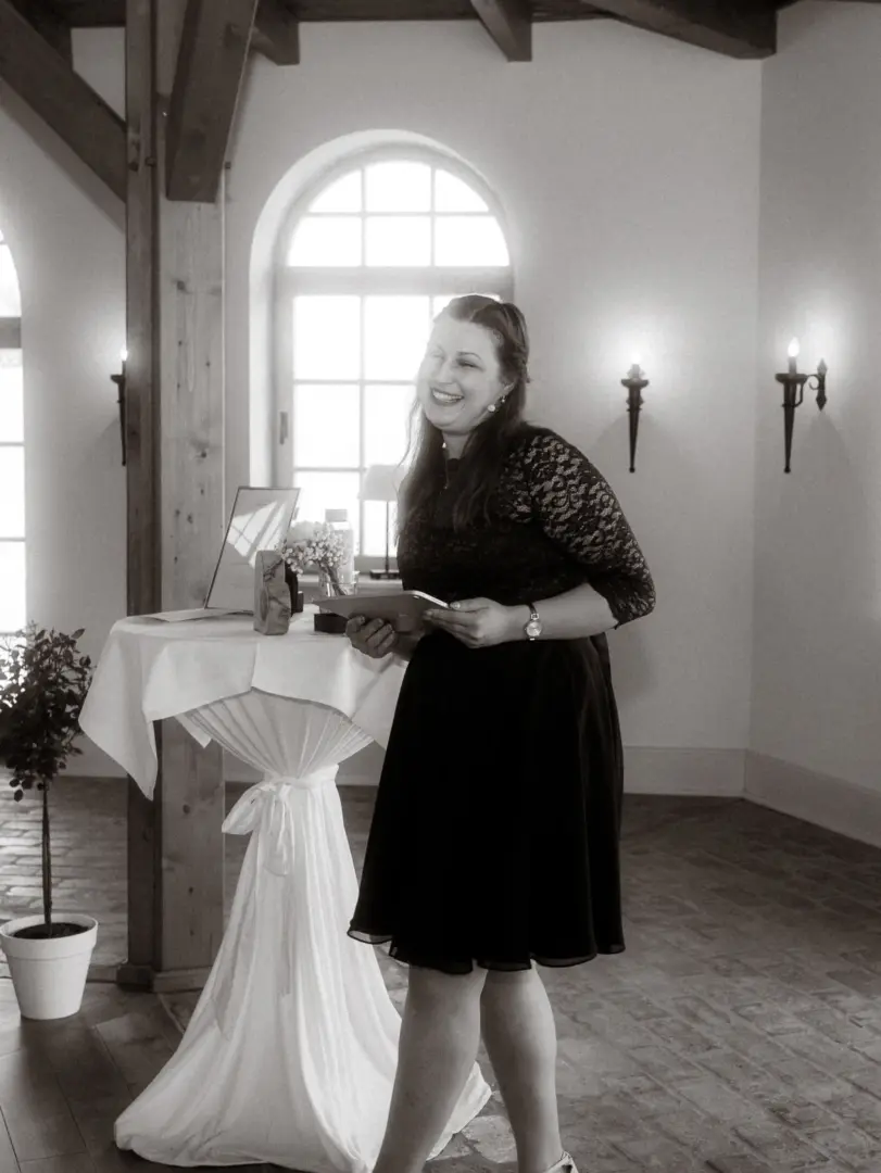 Eine fröhlich lachende Frau in einem eleganten schwarzen Spitzenkleid hält ein Tablet in den Händen und steht vor einem festlich gedeckten Stehtisch in einem hellen, rustikalen Raum. Professionelle Aufnahme von Ihrem Hochzeitsfotograf in Schwerin und MV.