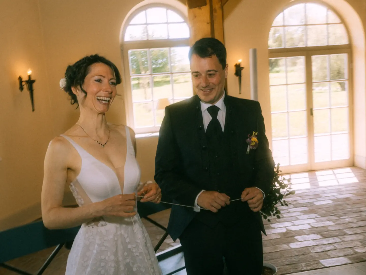 Ein glückliches Brautpaar bei einer symbolischen Zeremonie in einem hellen Raum mit großen Rundbogenfenstern. Die Braut lacht herzlich, während beide gemeinsam einen weißen Faden halten. Authentische Hochzeitsfotografie von Ihrem Hochzeitsfotograf in Schwerin und MV.