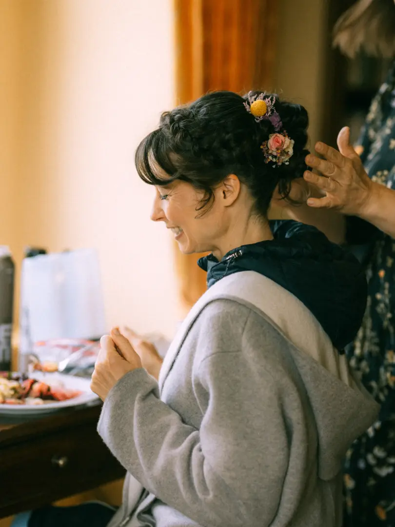Eine Braut mit geflochtener Hochsteckfrisur, die mit kleinen Blumen geschmückt ist, lächelt glücklich, während ihr Haar für die Hochzeit vorbereitet wird. Ein Hochzeitsfotograf in Schwerin und MV hält diesen intimen Moment der Vorbereitung fest.