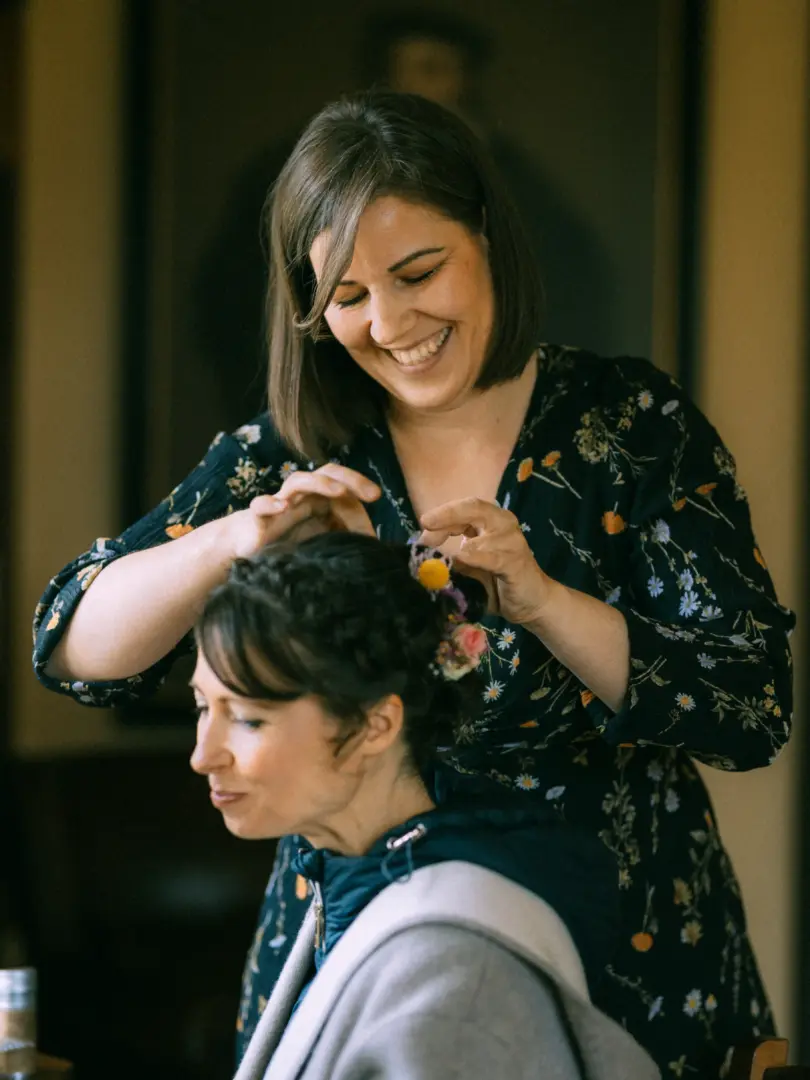 Eine freundliche Frau in einem dunkelblauen Kleid mit Blumenmuster steckt der Braut liebevoll kleine Blüten in die geflochtene Hochzeitsfrisur. Ein intimer Moment der Vorbereitung, festgehalten von Ihrem Hochzeitsfotograf in Schwerin und MV.