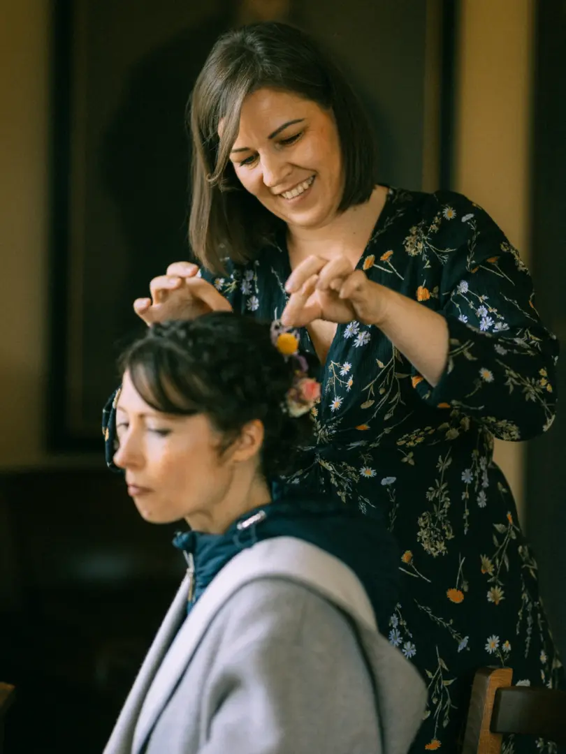Eine freundliche Stylistin steckt einer Braut mit geflochtenem Haar zarte Blumen in die Frisur. Die Braut trägt eine graue Kapuzenjacke, während die Stylistin ein dunkelblaues Kleid mit Blumenmuster trägt. Eine intime Vorbereitungsszene, festgehalten vom Hochzeitsfotograf in Schwerin und MV.