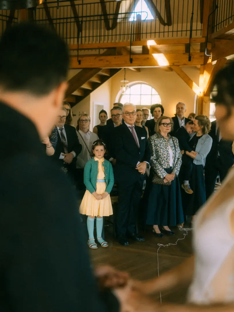 Ein Brautpaar hält sich während der Trauung an den Händen, während die Gäste in einer rustikalen Scheune andächtig zuschauen. Ein kleines Mädchen in einem gelben Kleid steht in der ersten Reihe. Professionelle Aufnahme von einem Hochzeitsfotograf in Schwerin und MV.