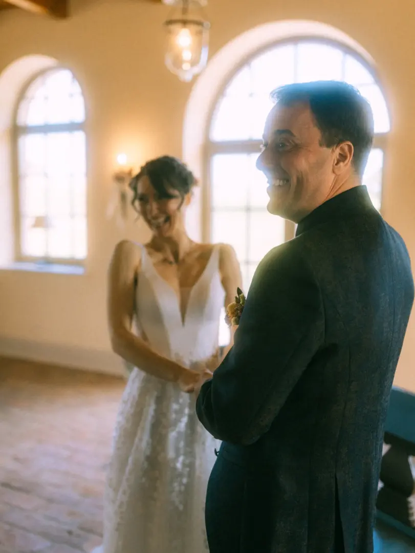 Ein glückliches Brautpaar hält sich bei einer emotionalen Zeremonie an den Händen. Der Bräutigam im dunklen Sakko lächelt seine Braut in einem eleganten weißen Hochzeitskleid strahlend an, während helles Licht durch die hohen Rundbogenfenster fällt. Ein Hochzeitsfotograf in Schwerin und MV.