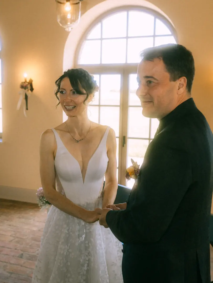 Ein glückliches Brautpaar hält sich während der Trauung an den Händen. Die Braut trägt ein weißes, tief ausgeschnittenes Spitzenkleid und lächelt ihren Partner liebevoll an, während der Bräutigam im dunklen Anzug vor einem hellen Fenster steht. Hochzeitsfotograf in Schwerin und MV.