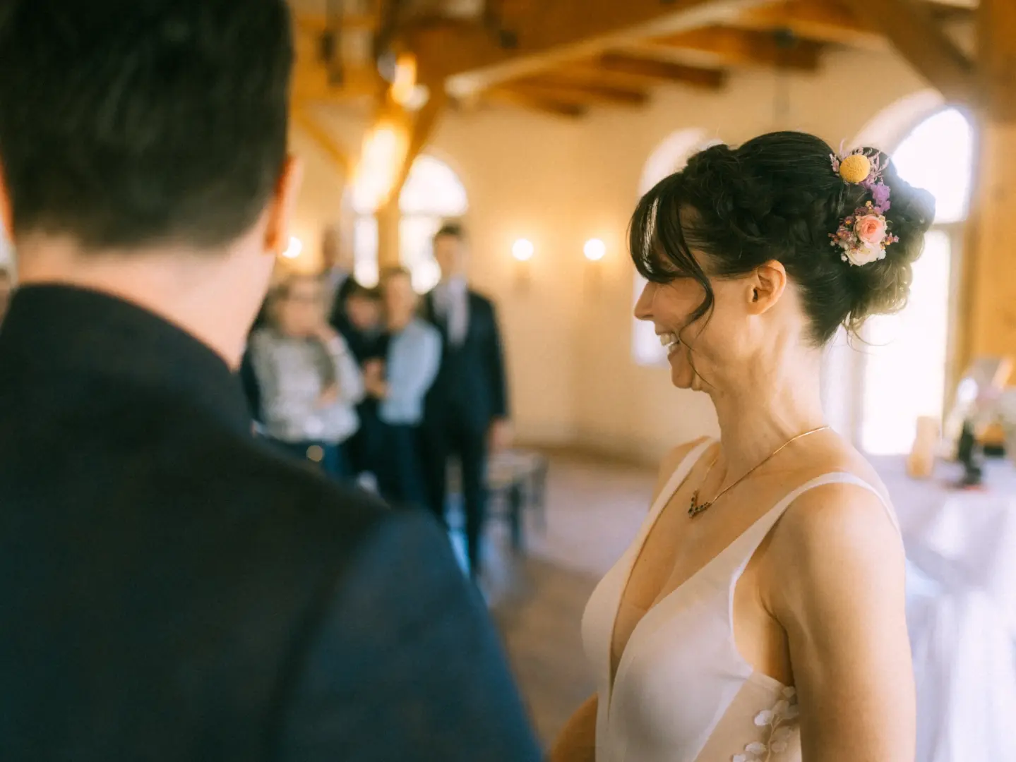 Eine glückliche Braut mit kunstvoller Hochsteckfrisur und Blumenschmuck lächelt ihrem Bräutigam während der Zeremonie liebevoll zu. Im Hintergrund sind unscharf die Hochzeitsgäste in einem hellen Saal zu sehen. Ein emotionaler Moment, festgehalten vom Hochzeitsfotograf in Schwerin und MV.