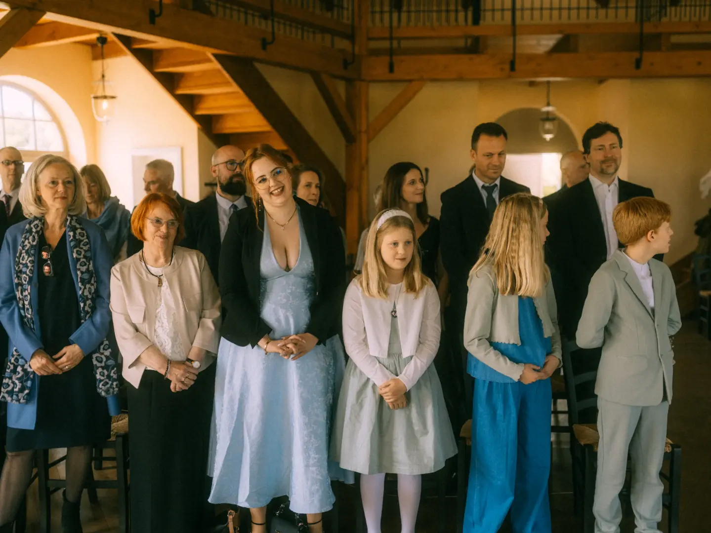 Eine festliche Hochzeitsgesellschaft steht in einem hellen, rustikalen Raum mit Holzbalken und verfolgt aufmerksam die Zeremonie. Die Gäste tragen elegante Kleidung in hellen und dunklen Tönen. Hochzeitsfotograf in Schwerin und MV.