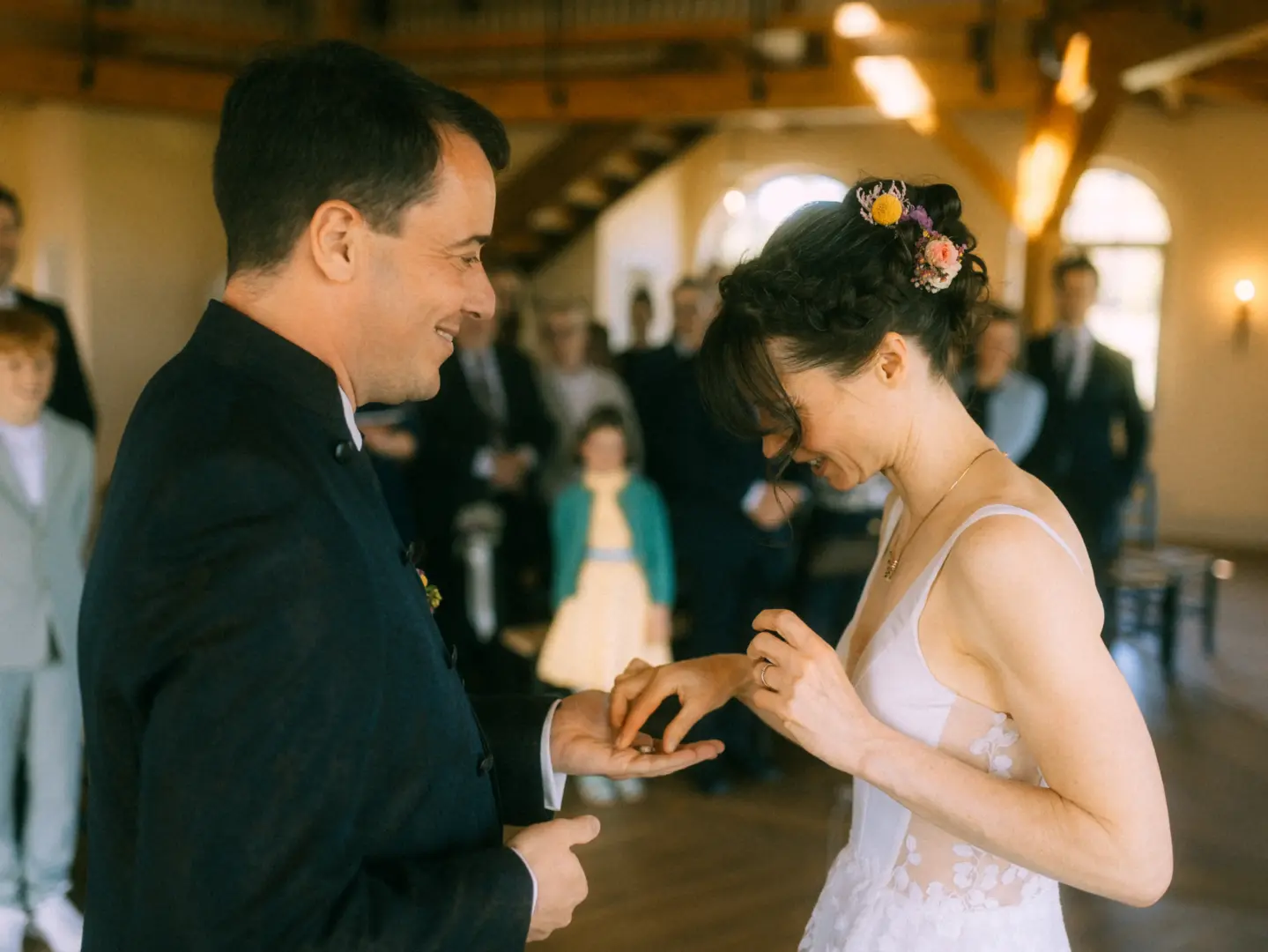 Ein Brautpaar während des Ringwechsels bei einer emotionalen Trauung: Die Braut mit Blumen im Haar legt dem Bräutigam im dunklen Anzug den Ehering an den Finger. Im Hintergrund sind unscharf Hochzeitsgäste zu sehen. Eine Aufnahme von Ihrem Hochzeitsfotograf in Schwerin und MV.