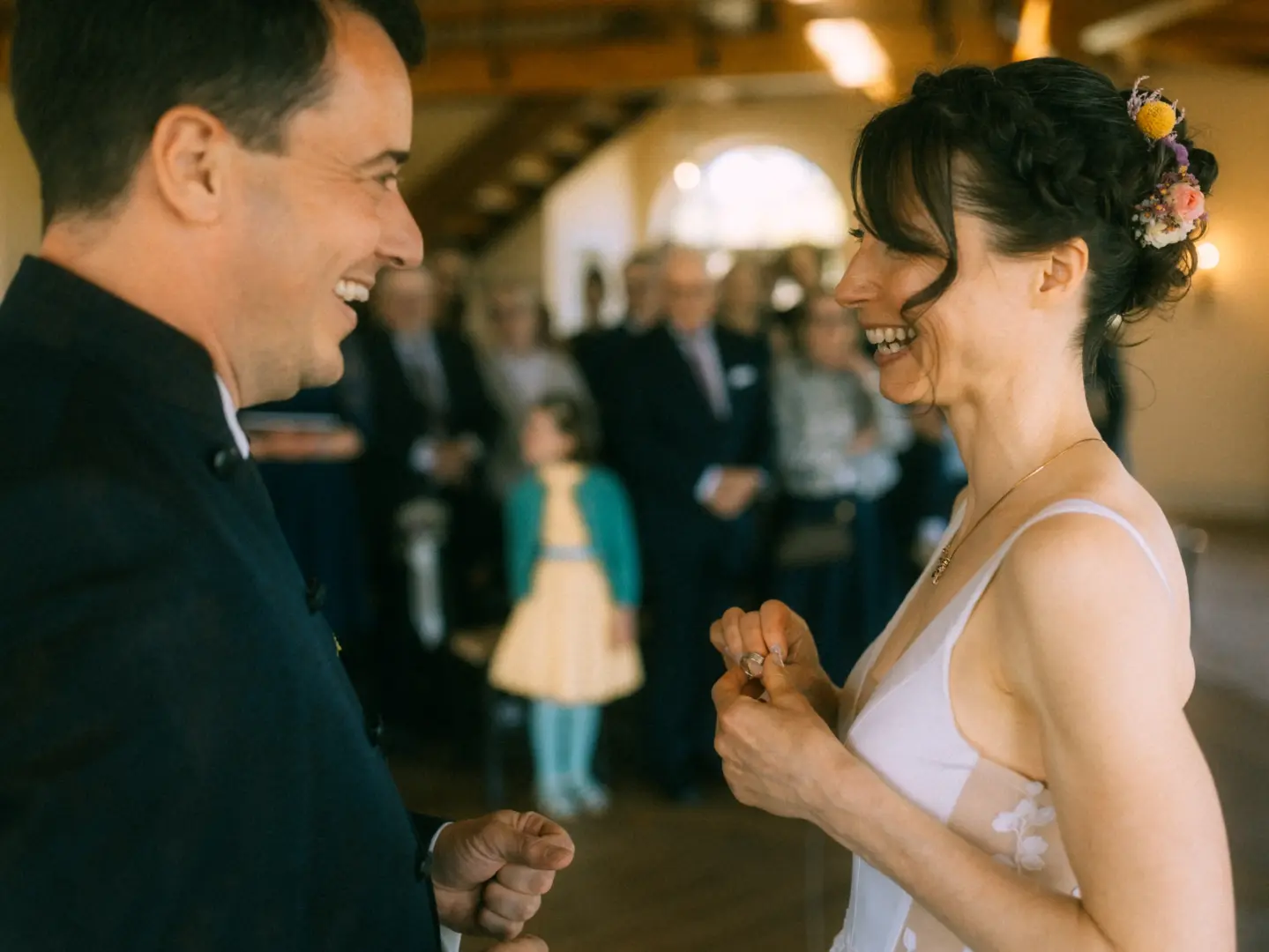 Ein glückliches Brautpaar lacht sich während der Trauung an, die Braut hält den Ehering in der Hand. Im Hintergrund sind unscharf die Hochzeitsgäste zu sehen. Eine authentische Aufnahme vom Hochzeitsfotograf in Schwerin und MV, die eine emotionale Lovestory einfängt.