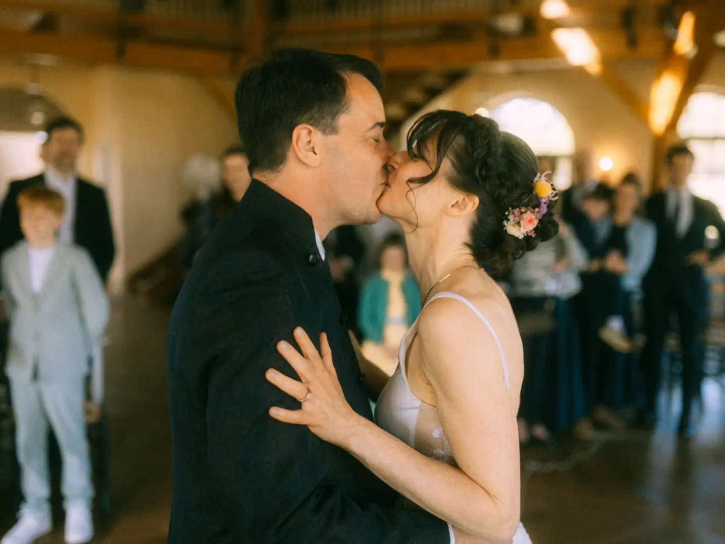 Ein Brautpaar küsst sich liebevoll bei einer Hochzeitsfeier in einer rustikalen Location. Die Braut trägt Blumen im Haar und ein elegantes Kleid, während der Bräutigam einen dunklen Anzug trägt. Lovestory Fotograf in Schwerin und MV für authentische Hochzeitsmomente.