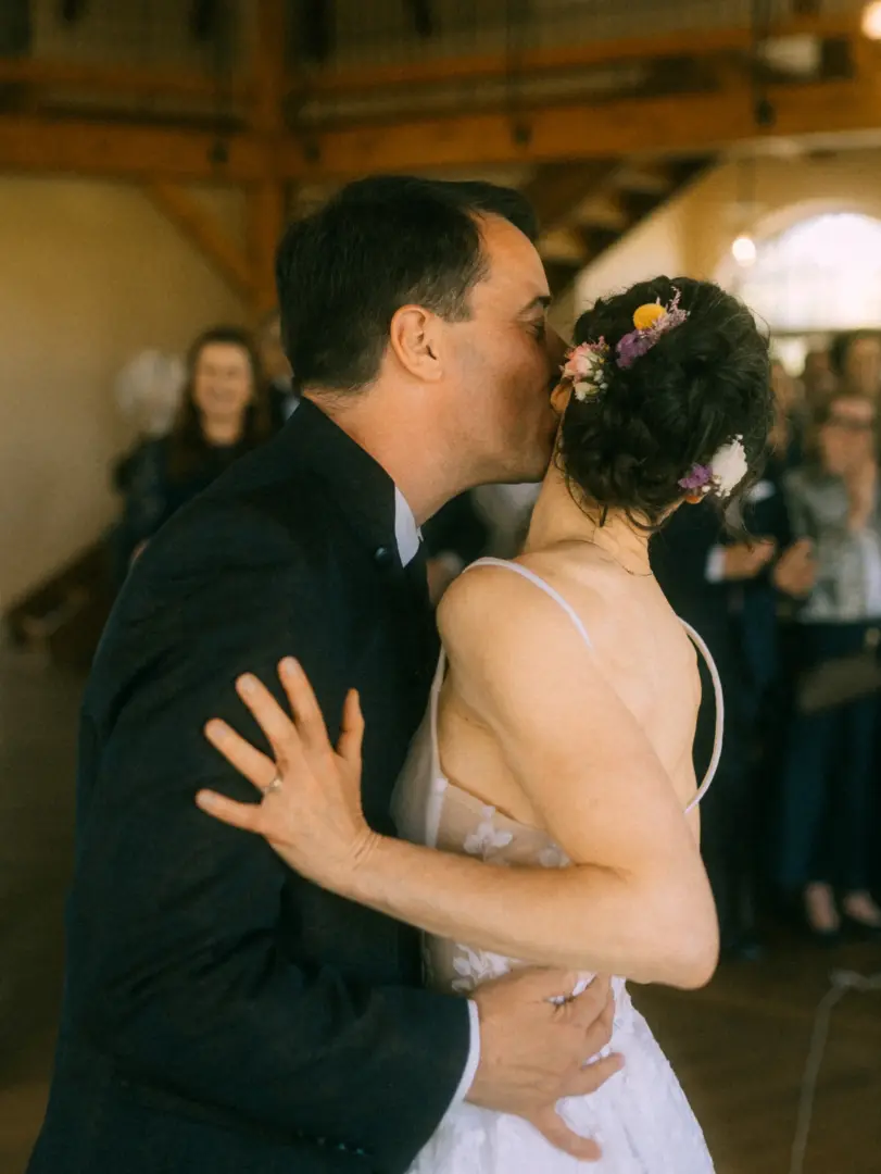 Ein Brautpaar in einer rustikalen Location während eines intimen Kusses. Die Braut trägt ein elegantes Hochzeitskleid mit Blumen im Haar, während der Bräutigam einen dunklen Anzug trägt. Eine einfühlsame Aufnahme von einem Lovestory Fotograf in Schwerin und MV.