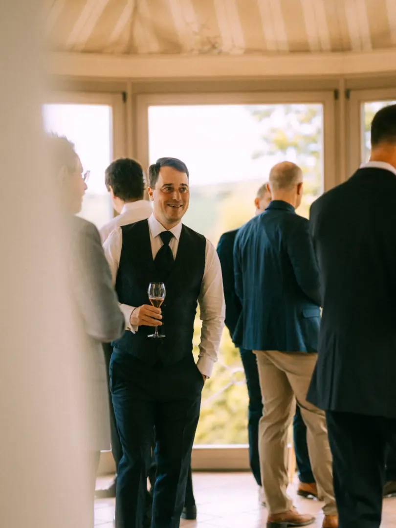 Ein lächelnder Gast in einem eleganten dunkelblauen Anzug mit Weste und Krawatte hält ein Weinglas in der Hand, während er sich unter andere Gäste mischt. Die Szene fängt die entspannte Stimmung bei einem Empfang ein, fotografiert von einem Hochzeitsfotograf in Schwerin und MV.