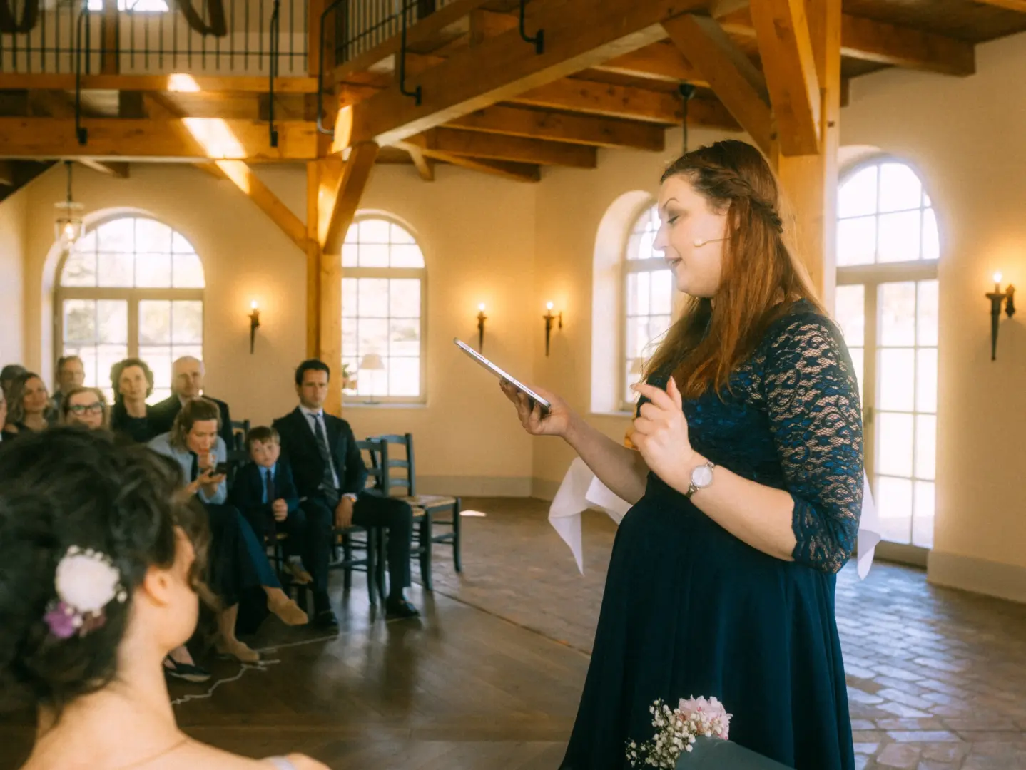 Eine Frau in einem dunkelblauen Spitzenkleid hält eine Rede bei einer Hochzeitszeremonie und liest von einem Tablet ab. Im Hintergrund sitzen Gäste in einem hellen, rustikalen Saal mit Holzbalken. Professioneller Hochzeitsfotograf in Schwerin und MV.