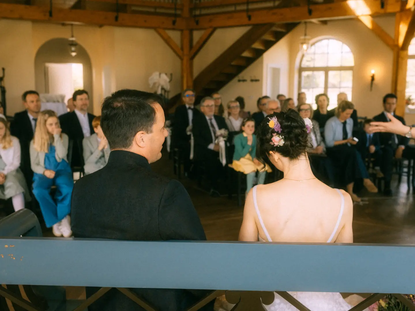 Brautpaar bei der Trauung in einer hellen, rustikalen Scheune, aufgenommen vom Hochzeitsfotograf in Schwerin und MV. Die Braut trägt Blumen im Haar, der Bräutigam einen dunklen Anzug. Im Hintergrund sitzen die Hochzeitsgäste in einer festlich geschmückten Location.