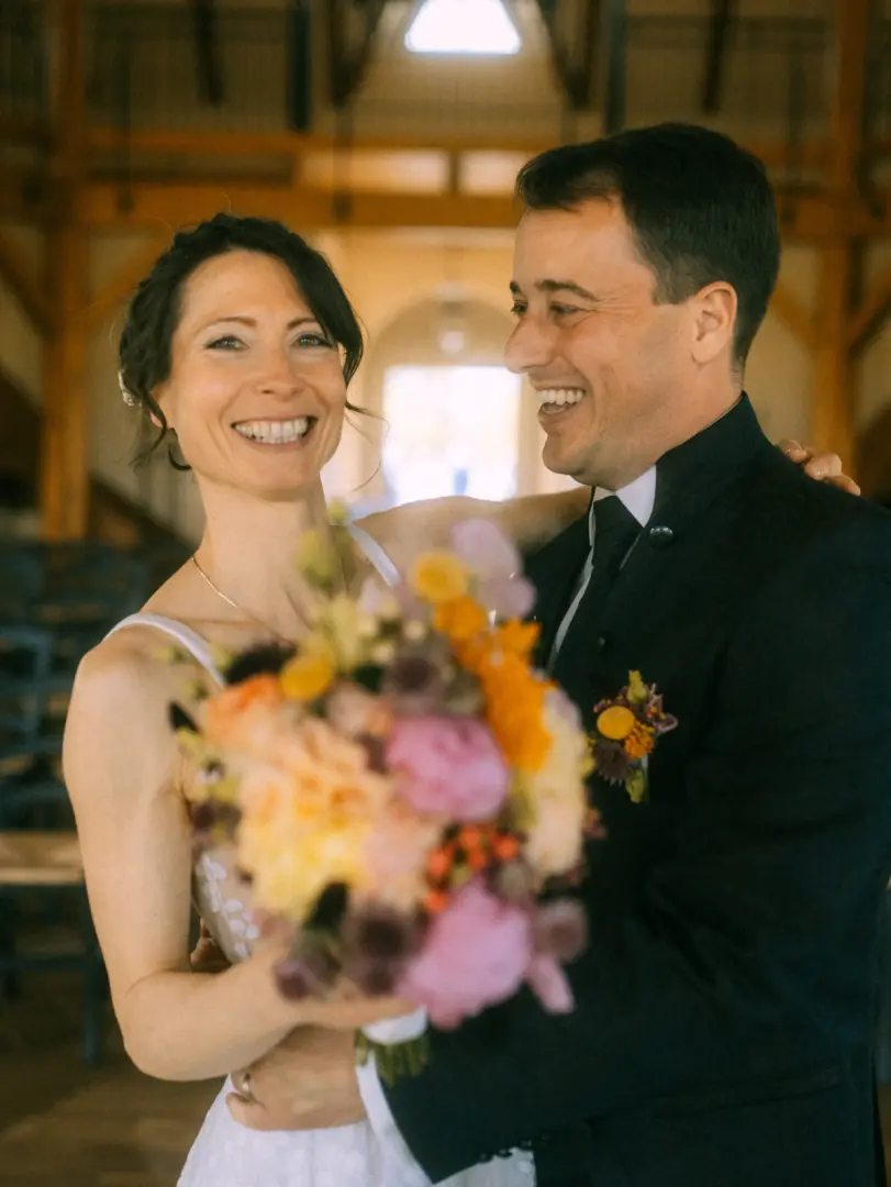 Ein glückliches Brautpaar lacht sich bei ihrer Hochzeit in einer rustikalen Scheune an. Die Braut hält einen bunten Blumenstrauß in den Vordergrund, während der Bräutigam im dunklen Anzug sie liebevoll im Arm hält. Eine authentische Aufnahme vom Lovestory Fotograf in Schwerin und MV.