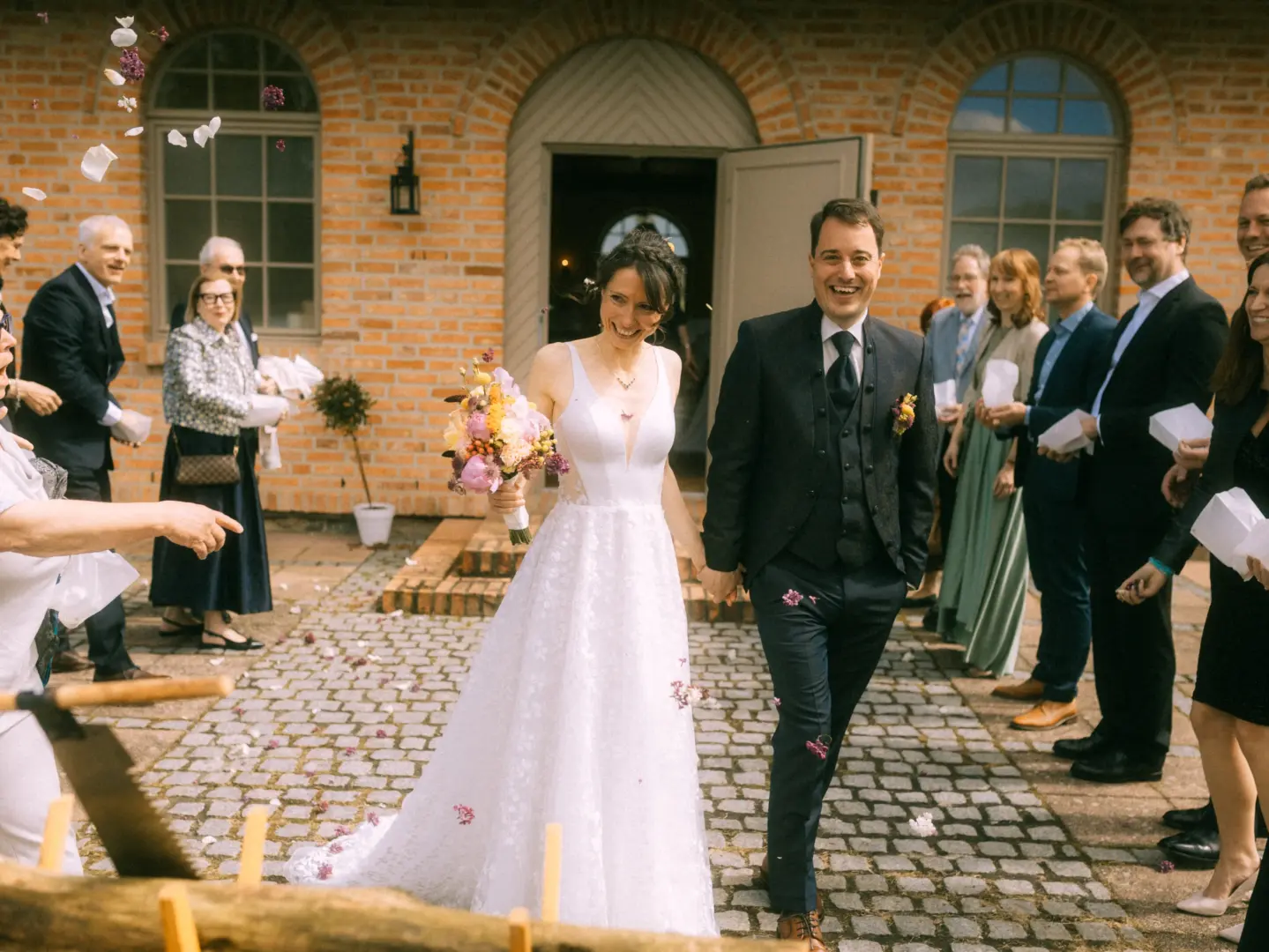 Ein glückliches Brautpaar verlässt nach der Trauung strahlend ein Backsteingebäude, während Gäste Blumen streuen. Die Braut trägt ein weißes Spitzenkleid und hält einen bunten Brautstrauß, der Bräutigam einen eleganten Anzug. Authentische Momente vom Hochzeitsfotograf in Schwerin und MV.