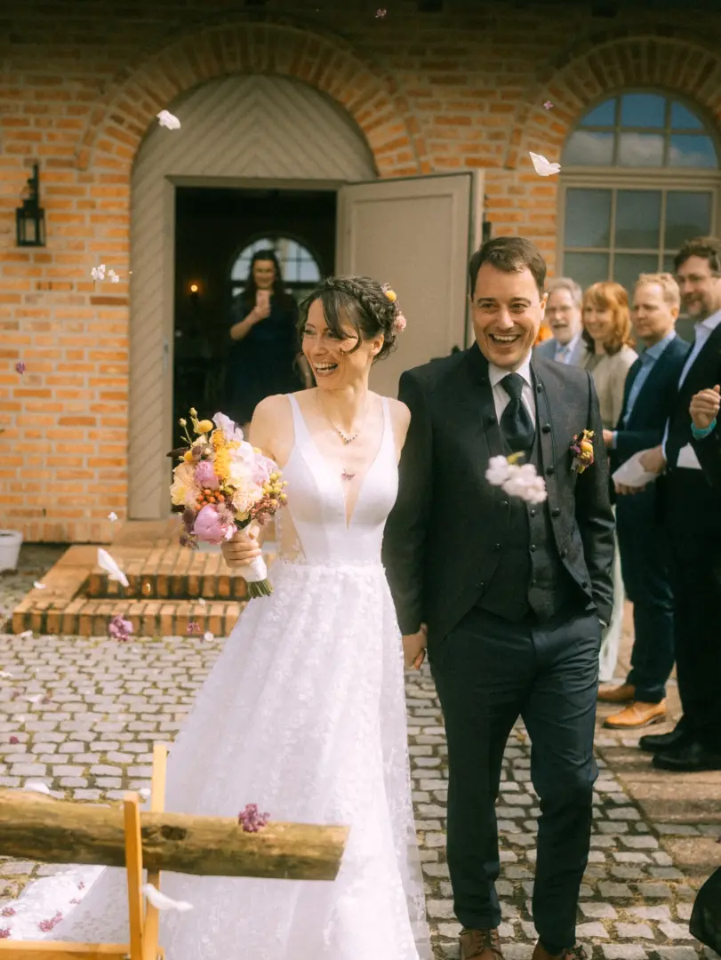Ein glückliches Brautpaar verlässt lachend das Standesamt, während Gäste Blumenblätter werfen. Die Braut trägt ein weißes Spitzenkleid und hält einen bunten Blumenstrauß, der Bräutigam einen eleganten Anzug. Ein stimmungsvoller Moment, festgehalten vom Hochzeitsfotograf in Schwerin und MV.