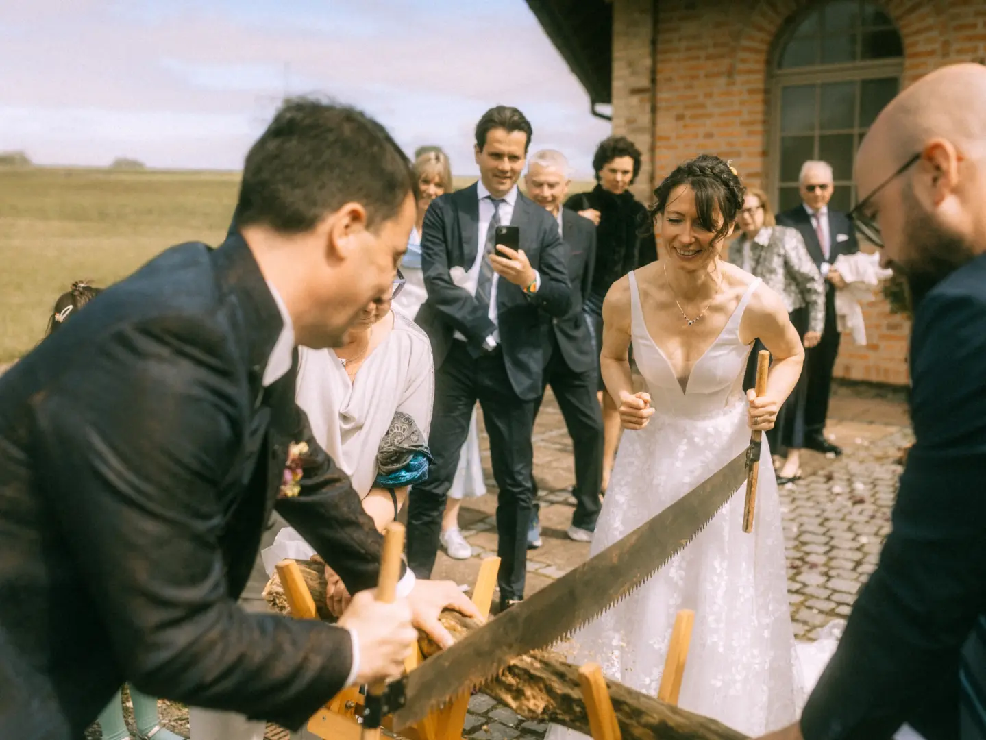 Brautpaar bei der traditionellen Holzstamm-Sägezeremonie während ihrer Hochzeit im Freien, beobachtet von lächelnden Gästen. Authentische Hochzeitsfotografie durch einen Hochzeitsfotograf in Schwerin und MV, die den freudigen Moment des gemeinsamen Sägens einfängt.