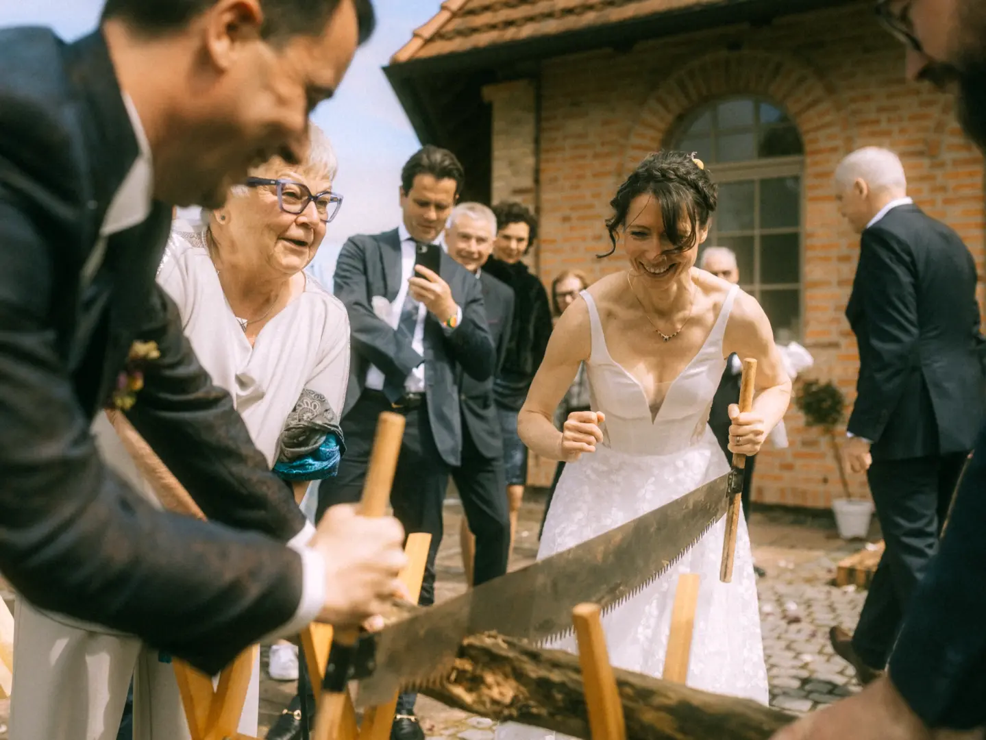 Ein Brautpaar sägt gemeinsam einen Baumstamm durch, eine traditionelle Hochzeitsaufgabe, während Gäste zuschauen und lächeln. Die Braut trägt ein weißes Kleid, der Bräutigam einen Anzug. Ein Hochzeitsfotograf in Schwerin und MV hält diesen freudigen Moment fest.