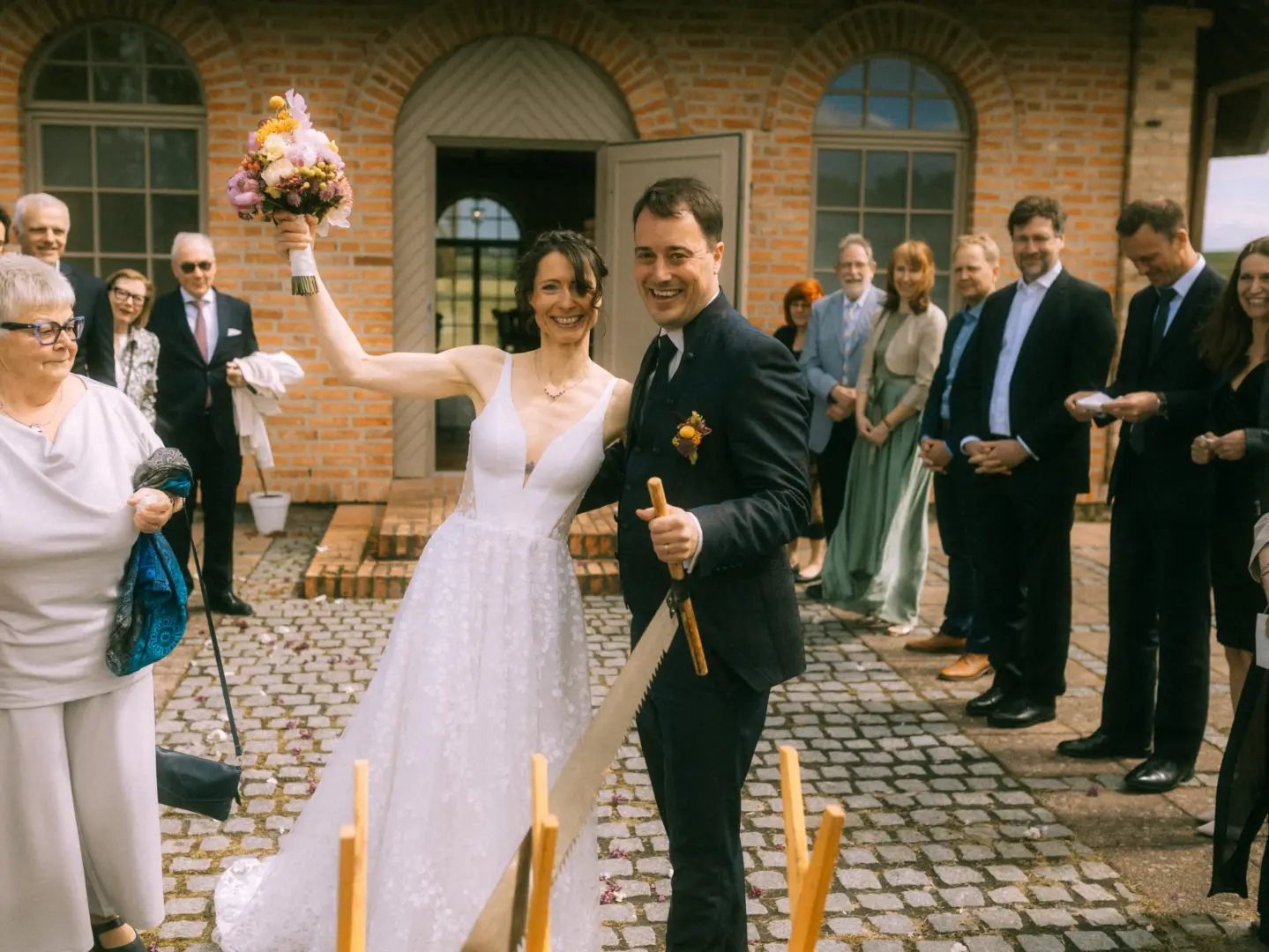 Ein glückliches Brautpaar beim traditionellen Baumstammsägen nach der Trauung vor einem historischen Backsteingebäude. Die Braut hält ihren Blumenstrauß freudig in die Luft, während Hochzeitsgäste im Hintergrund zuschauen. Hochzeitsfotograf in Schwerin und MV.