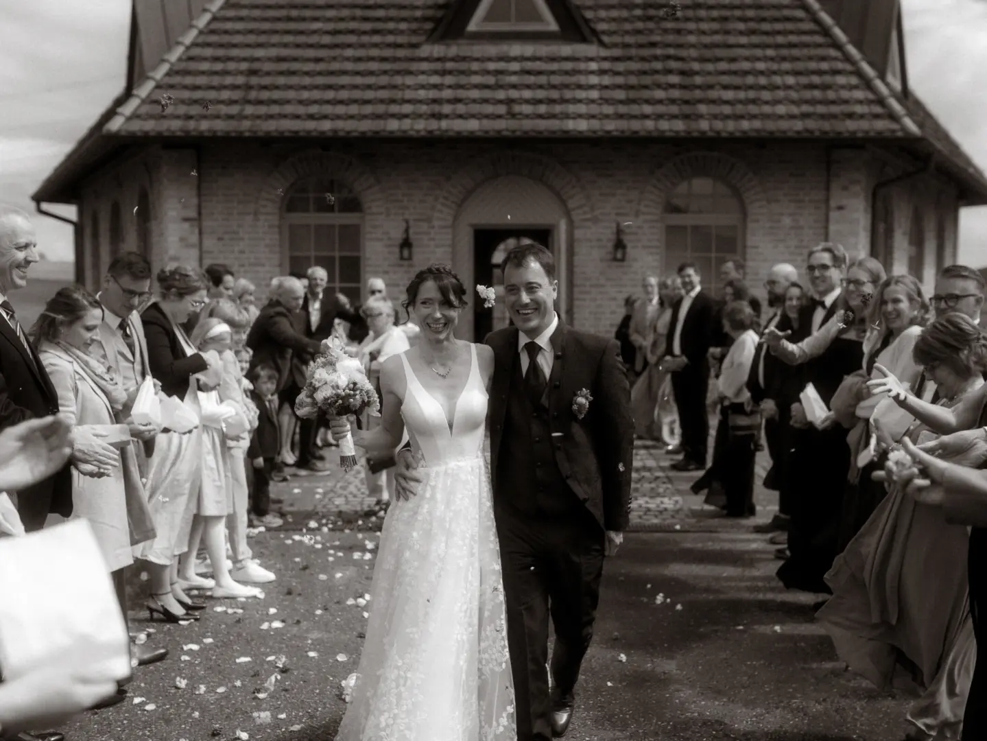 Ein glückliches Brautpaar schreitet nach der Trauung strahlend aus einem Backsteingebäude, während Gäste Blumen werfen. Diese authentische Momentaufnahme wurde von einem Hochzeitsfotograf in Schwerin und MV festgehalten, der die emotionale Atmosphäre perfekt einfängt.