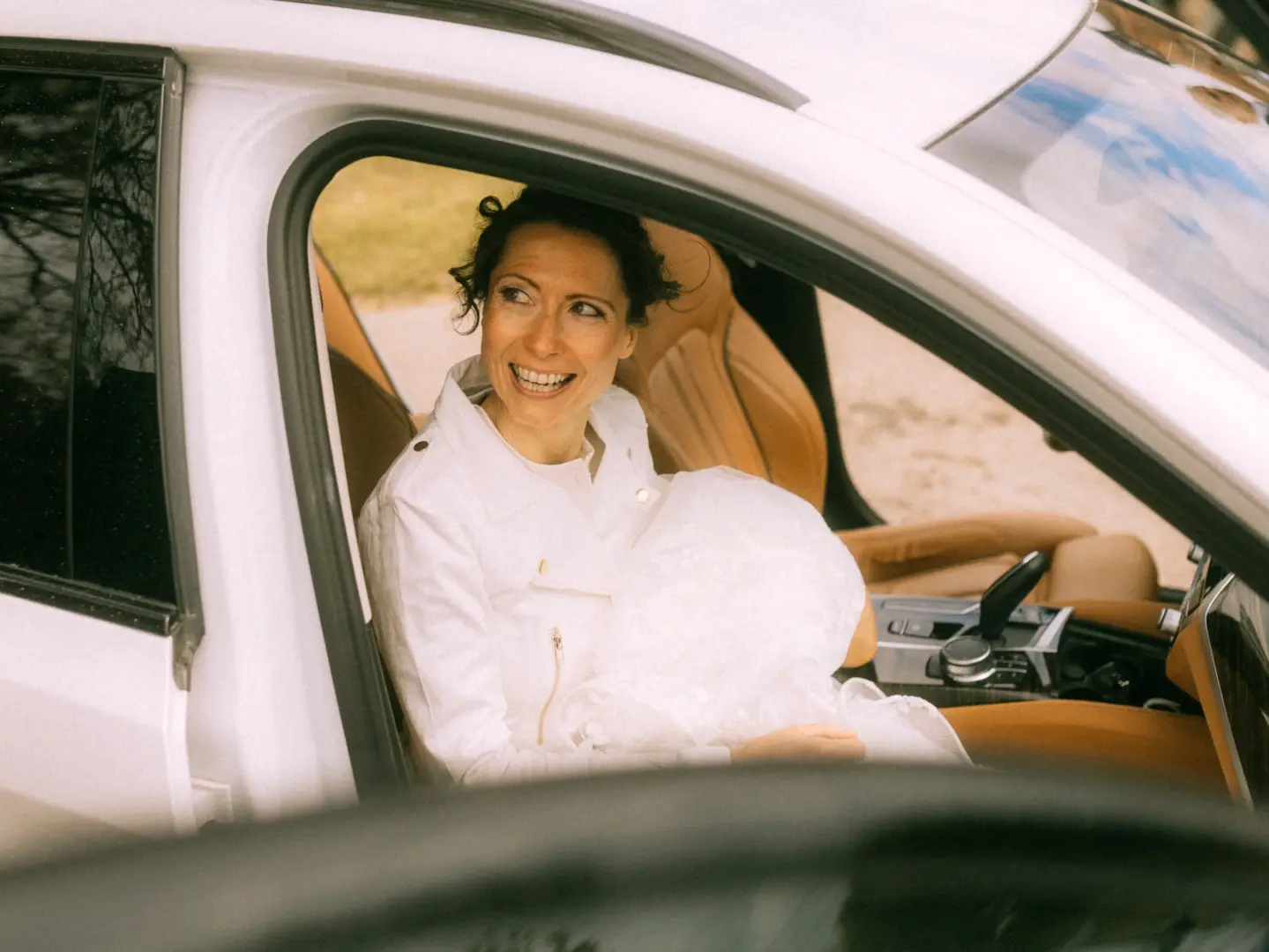 Eine strahlende Braut in einer weißen Jacke sitzt auf dem Beifahrersitz eines Autos und blickt glücklich zur Seite. Das Foto fängt einen besonderen Moment ein, wie er für einen Hochzeitsfotograf in Schwerin und MV typisch ist.