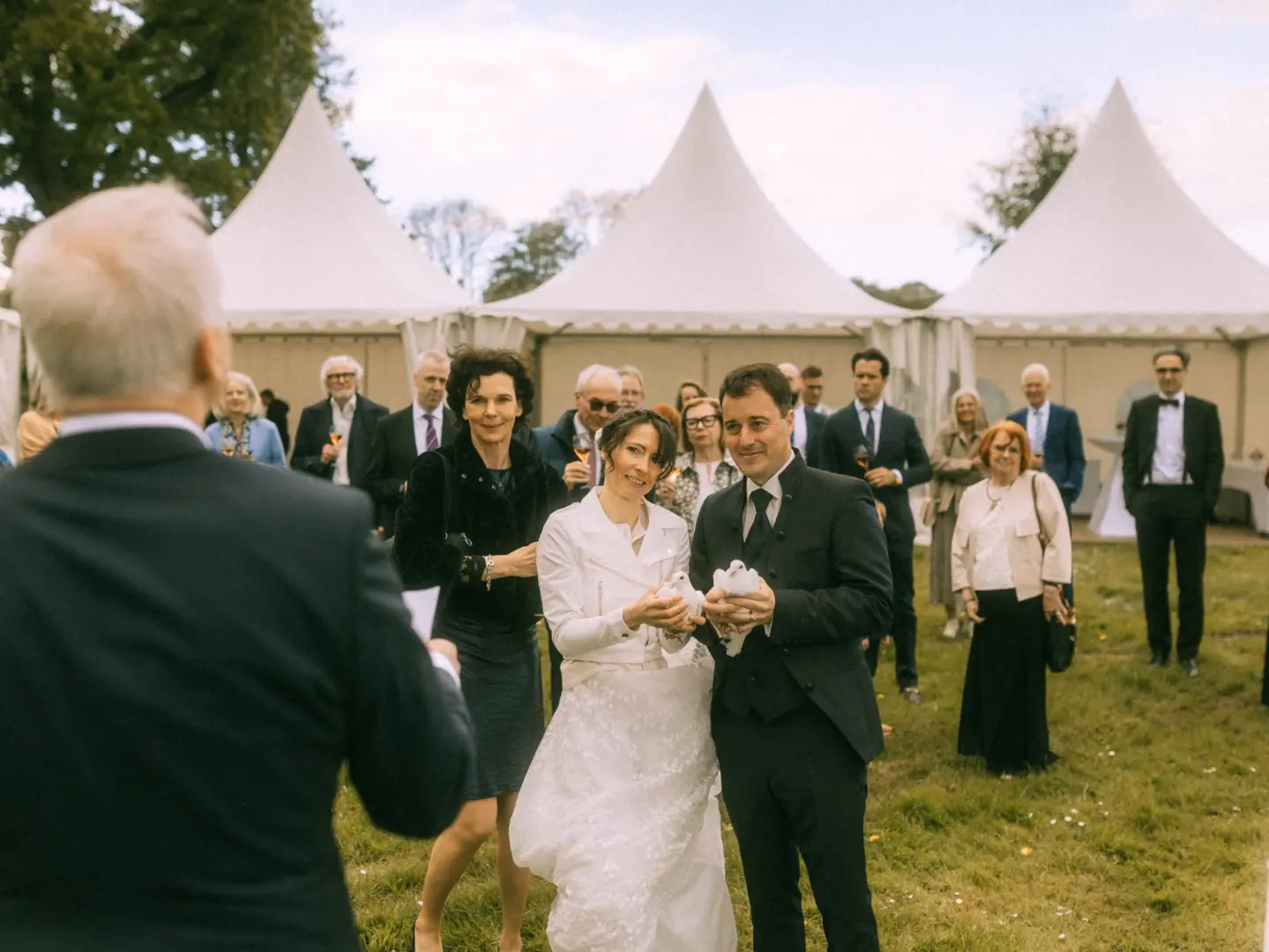 Ein Brautpaar hält bei einer Hochzeitszeremonie im Freien weiße Tauben in den Händen, umgeben von Gästen vor weißen Festzelten. Diese emotionale Aufnahme wurde von einem Hochzeitsfotograf in Schwerin und MV festgehalten.
