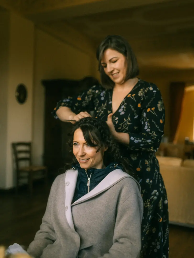 Eine lächelnde Braut sitzt in einer beigen Strickjacke, während ihr eine Stylistin in einem dunkelblauen Blumenkleid die Haare für den großen Tag frisiert. Ein intimer Moment der Vorbereitung, festgehalten von Ihrem Hochzeitsfotograf in Schwerin und MV.