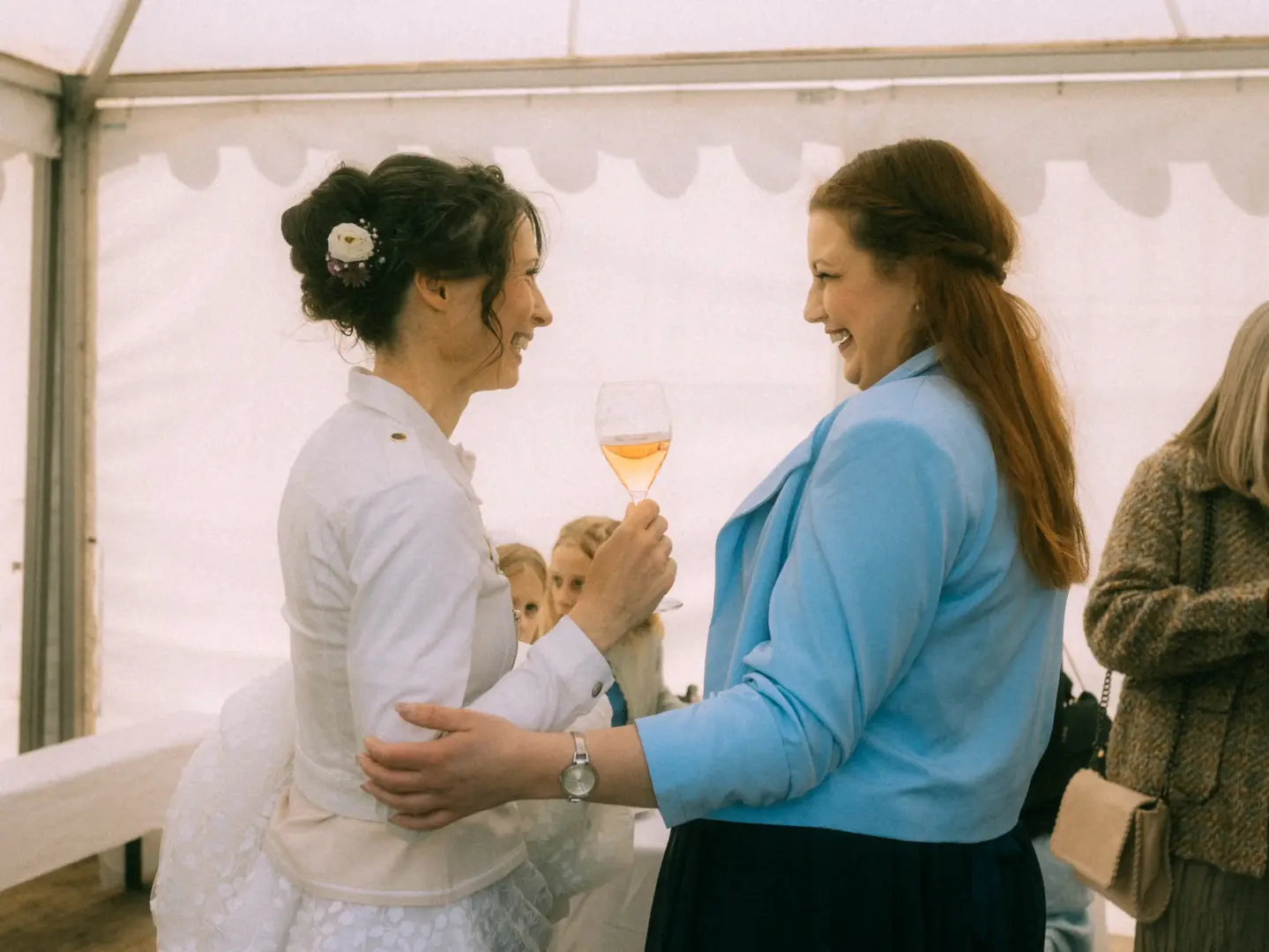 Zwei fröhliche Frauen stoßen in einem hellen Festzelt mit einem Glas Roséwein an. Die Braut trägt eine weiße Jacke über ihrem Brautkleid, während ihre Begleiterin in einem blauen Blazer strahlt. Ein stimmungsvoller Moment, eingefangen von einem Hochzeitsfotograf in Schwerin und MV.