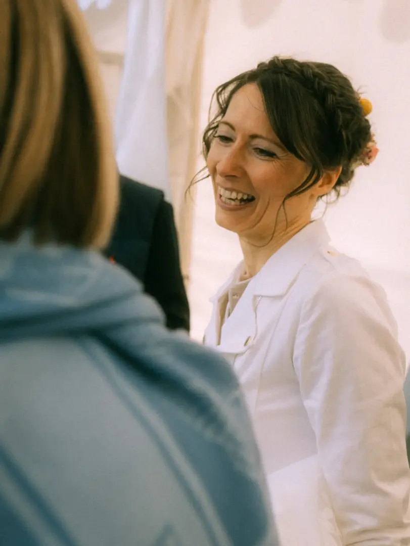 Eine strahlende Braut mit geflochtener Hochsteckfrisur und weißem Blazer lacht herzlich während eines Gesprächs auf ihrer Hochzeitsfeier. Diese authentische Aufnahme wurde von einem Hochzeitsfotograf in Schwerin und MV festgehalten.