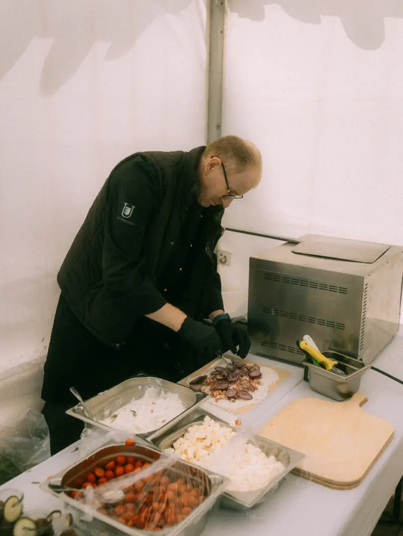 Ein Koch in schwarzer Weste mit dem Logo „Diemshusen“ bereitet konzentriert einen Wrap oder Flammkuchen zu. Er belegt den Teig mit roten Zwiebeln, während vor ihm in Edelstahlschalen frische Zutaten wie Tomaten und Käsewürfel für das Catering bereitstehen.
