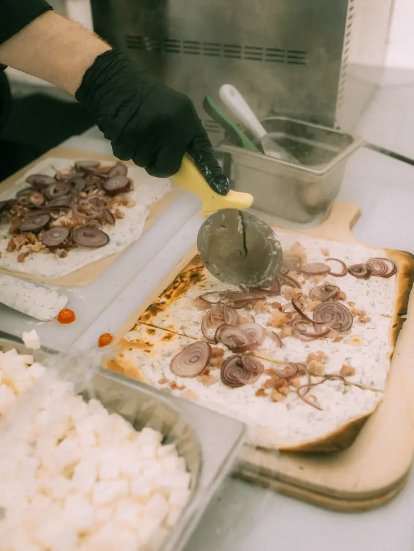 Ein Koch mit schwarzen Handschuhen schneidet eine frisch belegte Flammkuchen-Spezialität mit roten Zwiebeln und Speck auf einem Holzbrett. Im Vordergrund befinden sich Behälter mit gewürfeltem Käse, während im Hintergrund weiteres Equipment für die professionelle Zubereitung bereitsteht.
