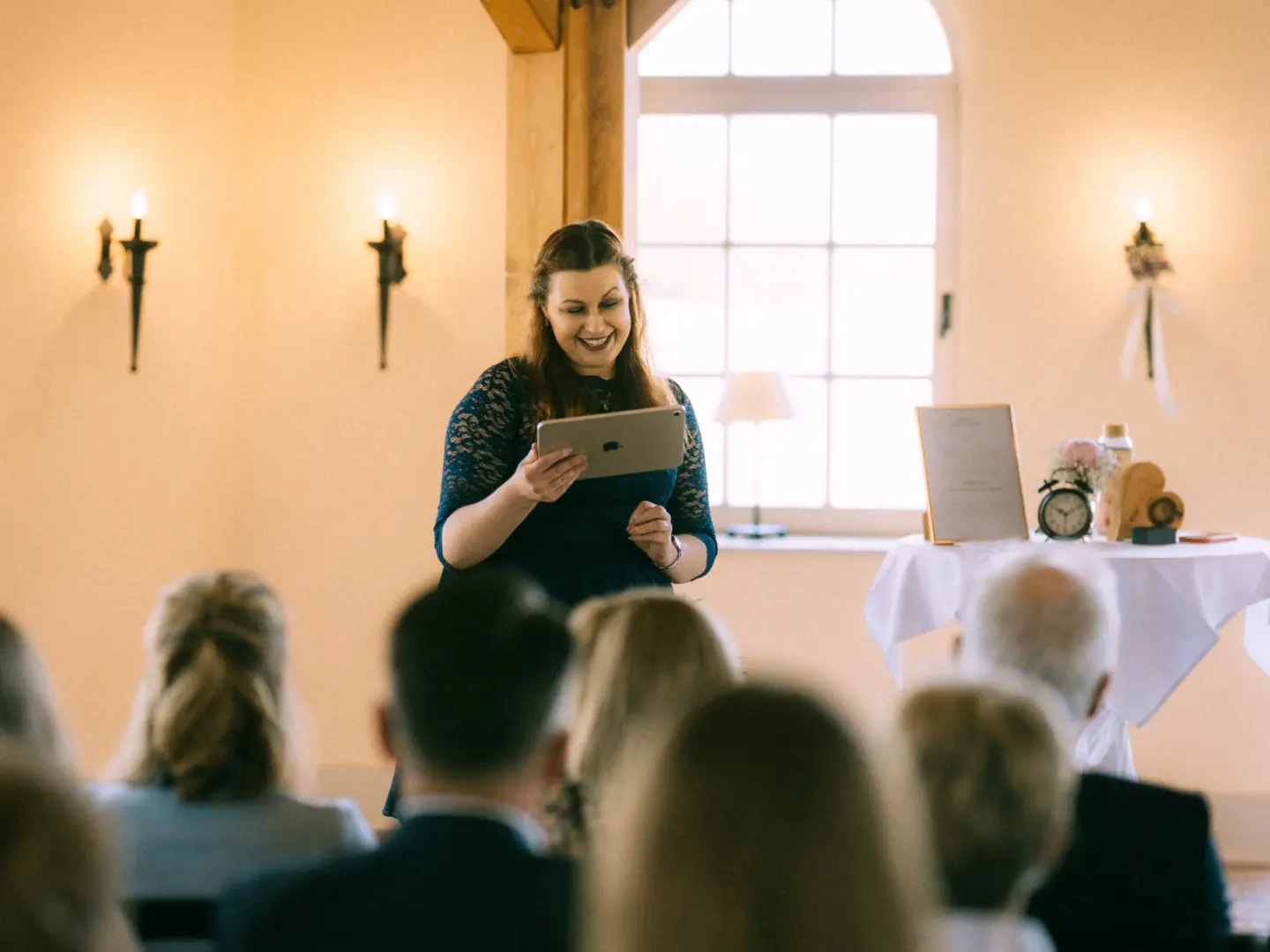 Eine lächelnde Frau in einem dunkelblauen Spitzenkleid hält bei einer Hochzeitszeremonie ein Tablet in den Händen und trägt eine Rede vor. Im Hintergrund ist ein helles Fenster mit Tageslicht zu sehen. Hochzeitsfotograf in Schwerin und MV.
