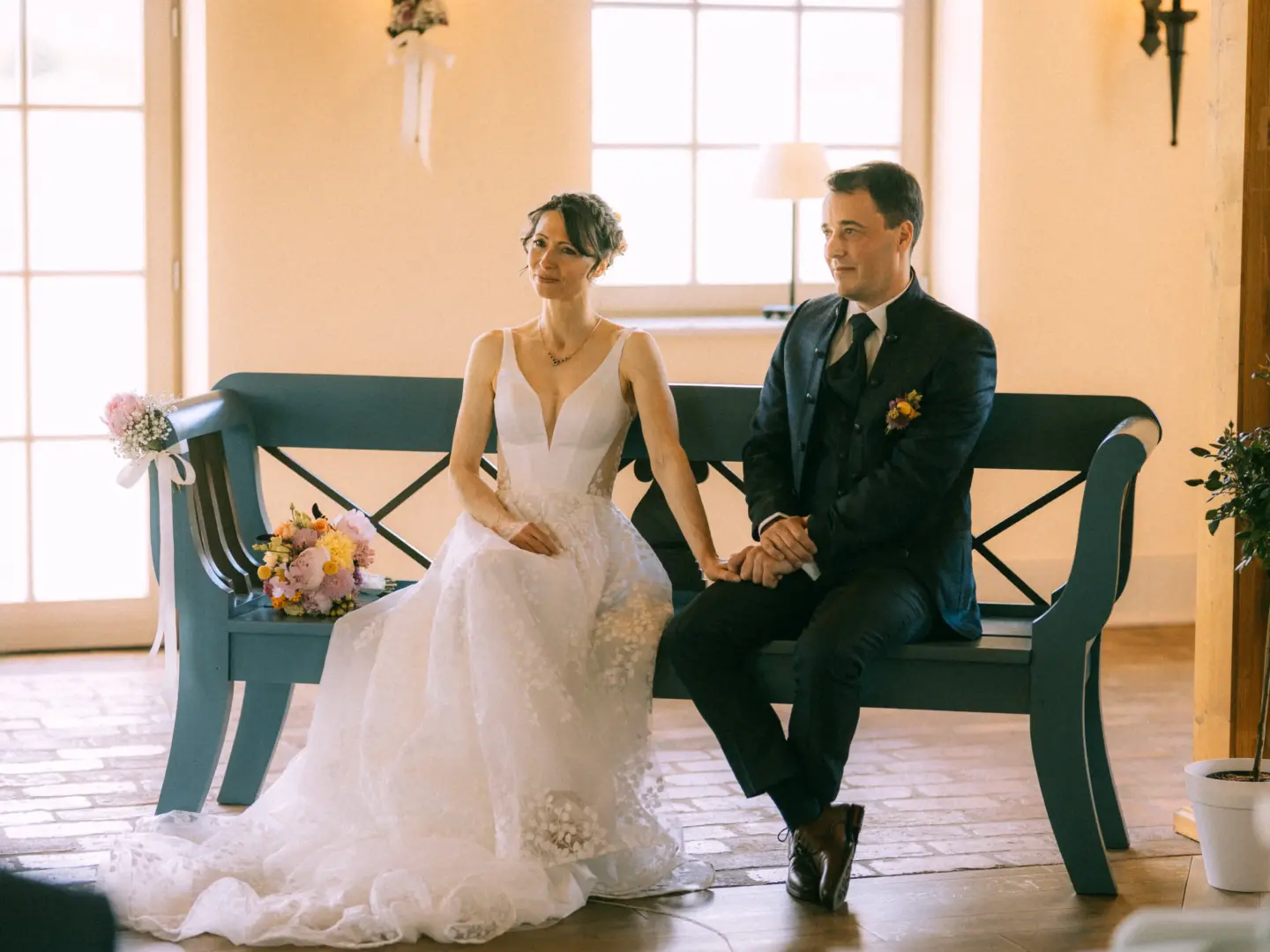 Ein Brautpaar sitzt während der Trauung auf einer blauen Holzbank. Die Braut trägt ein weißes, besticktes Hochzeitskleid, der Bräutigam einen eleganten dunklen Anzug. Die Szene wirkt emotional und festlich. Lovestory Fotograf in Schwerin und MV.