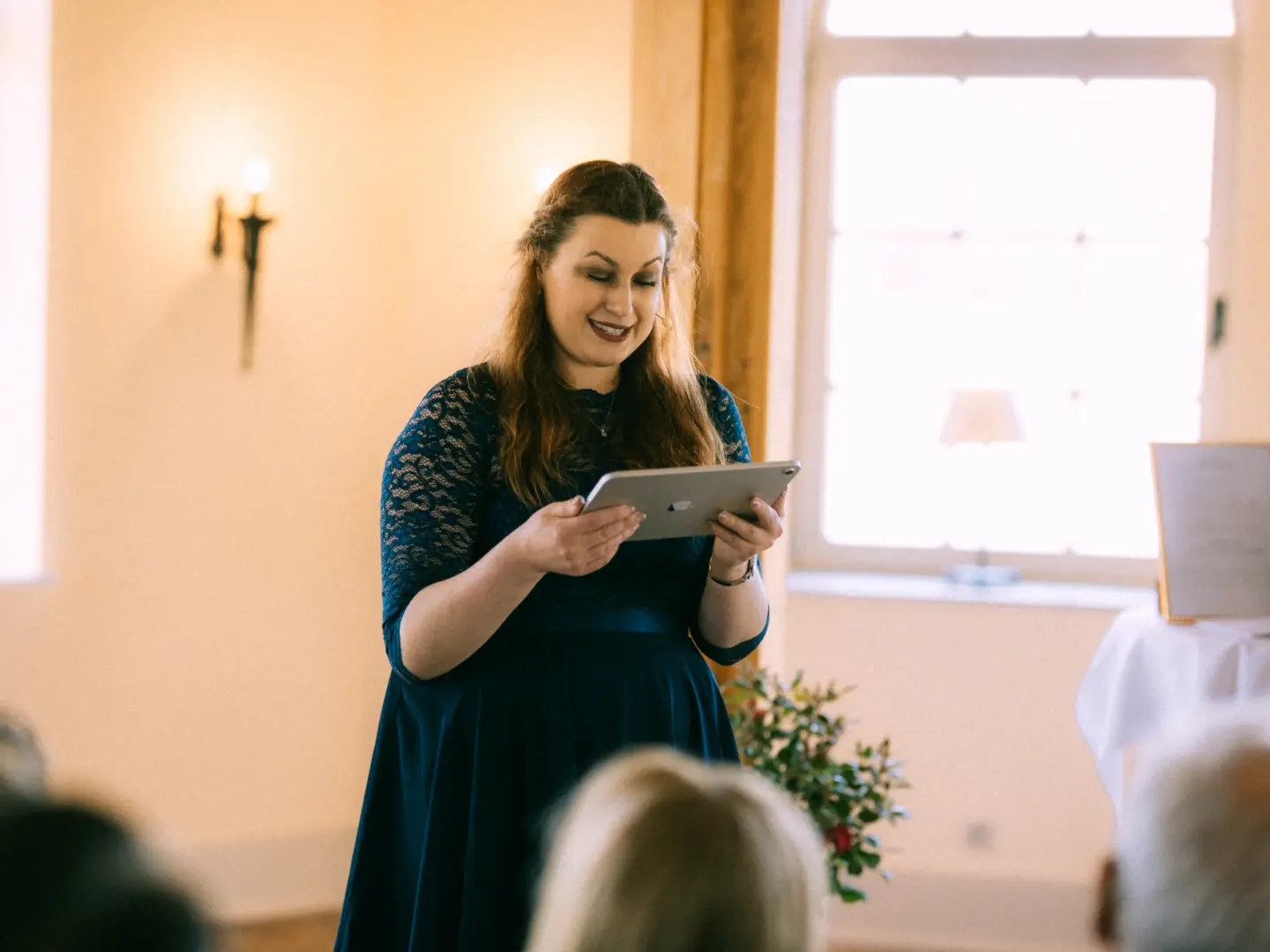 Eine lächelnde Frau in einem eleganten dunkelblauen Spitzenkleid hält während einer Hochzeitszeremonie ein Tablet in den Händen und trägt eine Rede vor. Im Hintergrund ist ein heller Raum mit einem Fenster zu sehen. Hochzeitsfotograf in Schwerin und MV.
