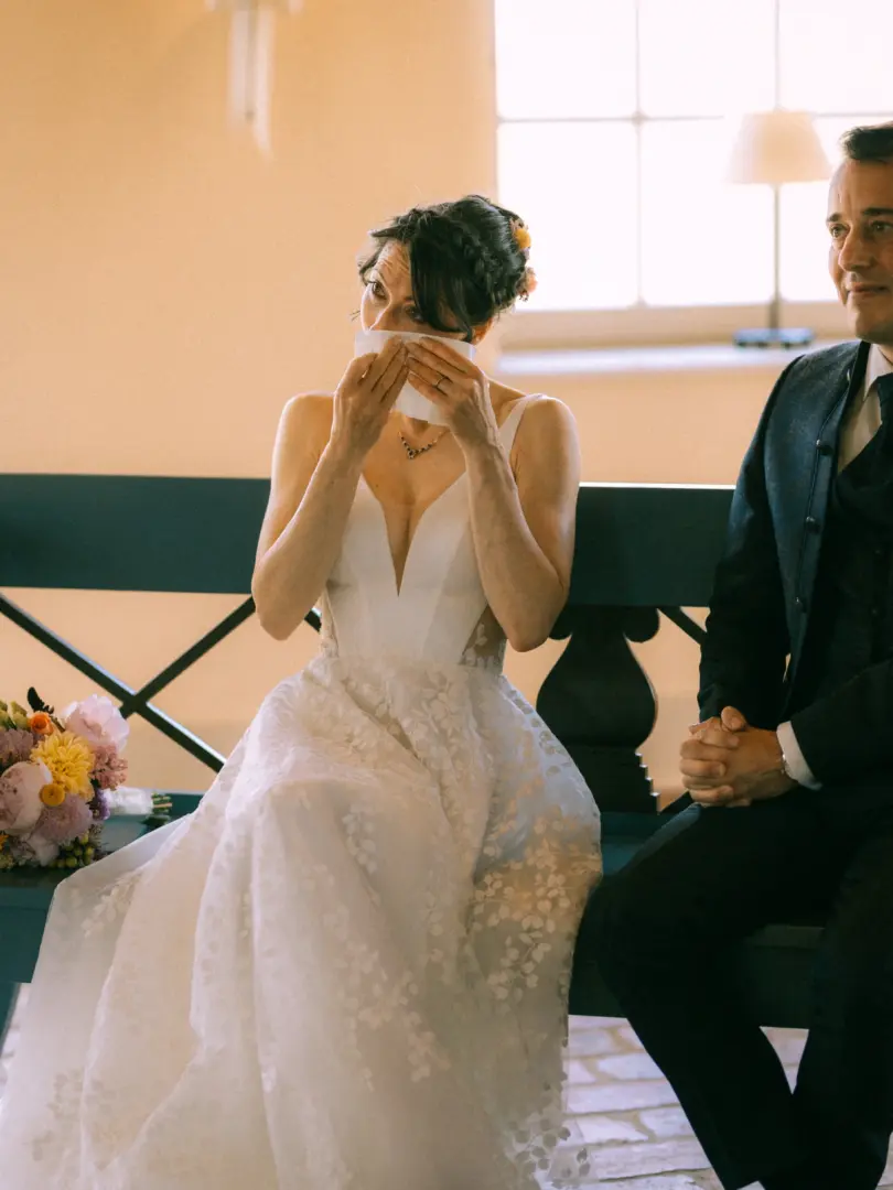 Eine Braut in einem weißen Spitzenkleid wischt sich gerührt eine Träne mit einem Taschentuch ab, während sie neben ihrem Bräutigam auf einer Bank sitzt. Ein romantischer Moment, eingefangen von einem Hochzeitsfotograf in Schwerin und MV.