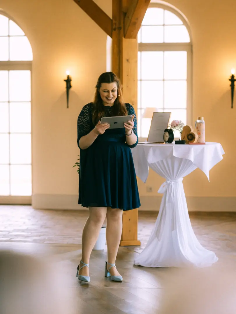 Eine lächelnde Frau in einem dunkelblauen Spitzenkleid hält ein Tablet in den Händen und steht vor einem mit weißem Tuch gedeckten Stehtisch in einem hellen, eleganten Saal. Diese Aufnahme zeigt die Arbeit als Hochzeitsfotograf in Schwerin und MV bei einer feierlichen Zeremonie.