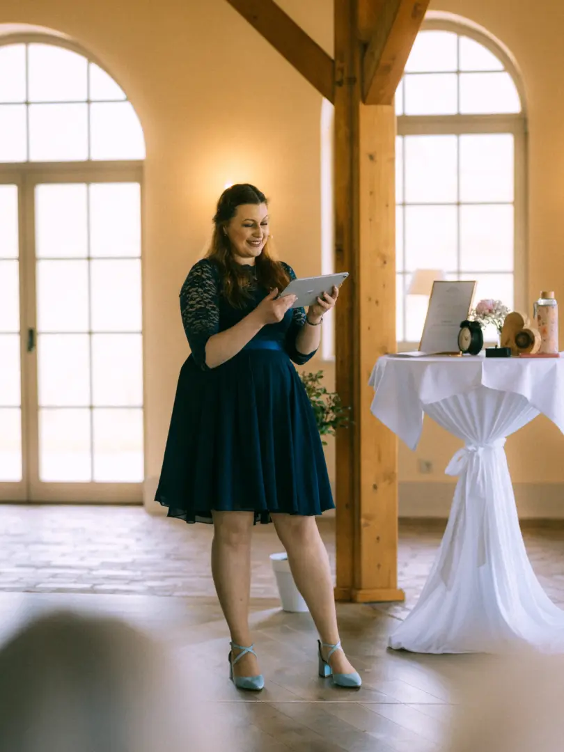Eine junge Frau in einem eleganten dunkelblauen Spitzenkleid hält lächelnd ein Tablet in den Händen und hält eine Rede bei einer Hochzeitsfeier. Im Hintergrund steht ein mit weißem Stoff bedeckter Stehtisch in einem hellen Raum. Professioneller Hochzeitsfotograf in Schwerin und MV.