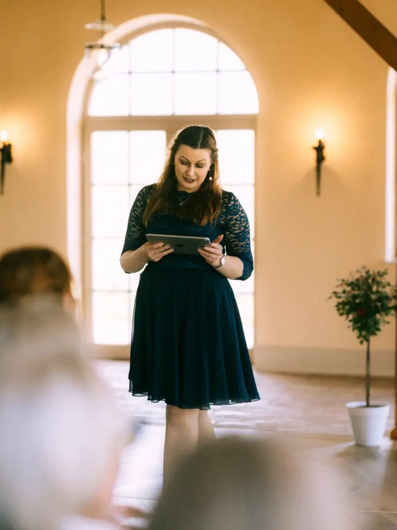 Eine Frau mit langem braunem Haar trägt ein dunkelblaues Spitzenkleid und hält ein Tablet in den Händen, während sie bei einer Trauung spricht. Im Hintergrund ist ein heller Saal mit großen Fenstern zu sehen. Hochzeitsfotograf in Schwerin und MV.