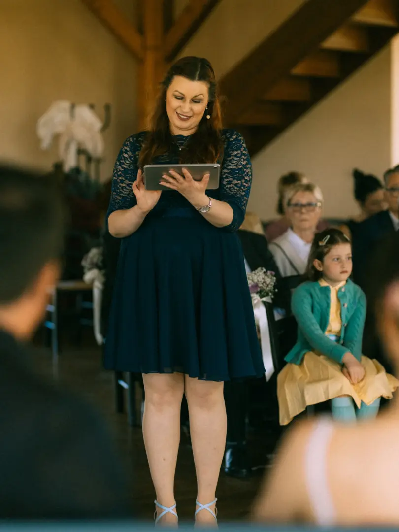 Eine lächelnde Frau in einem dunkelblauen Spitzenkleid hält während einer Hochzeitszeremonie ein Tablet in den Händen und trägt ihre Rede vor. Im Hintergrund sitzen Gäste, darunter ein kleines Mädchen in einem gelben Kleid. Hochzeitsfotograf in Schwerin und MV.