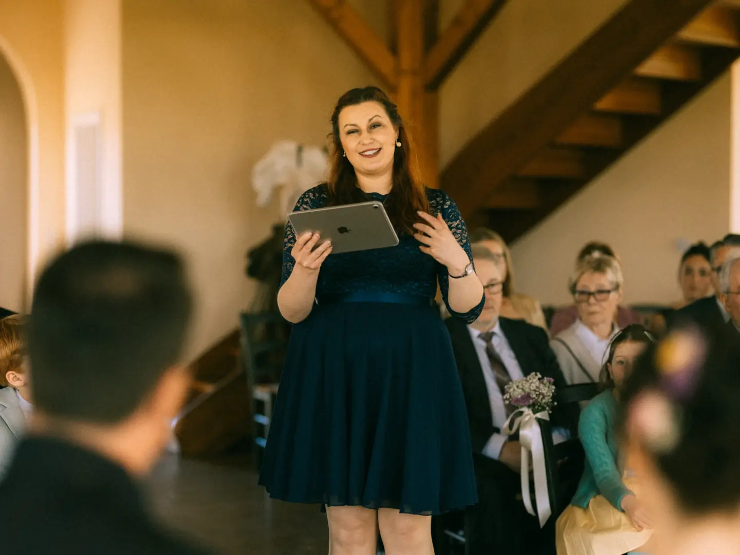 Eine Frau in einem dunkelblauen Spitzenkleid hält während einer Hochzeitsrede ein Tablet in den Händen und spricht zu den Gästen. Die festliche Atmosphäre wird durch den Hochzeitsfotograf in Schwerin und MV festgehalten, während das Publikum aufmerksam zuhört.