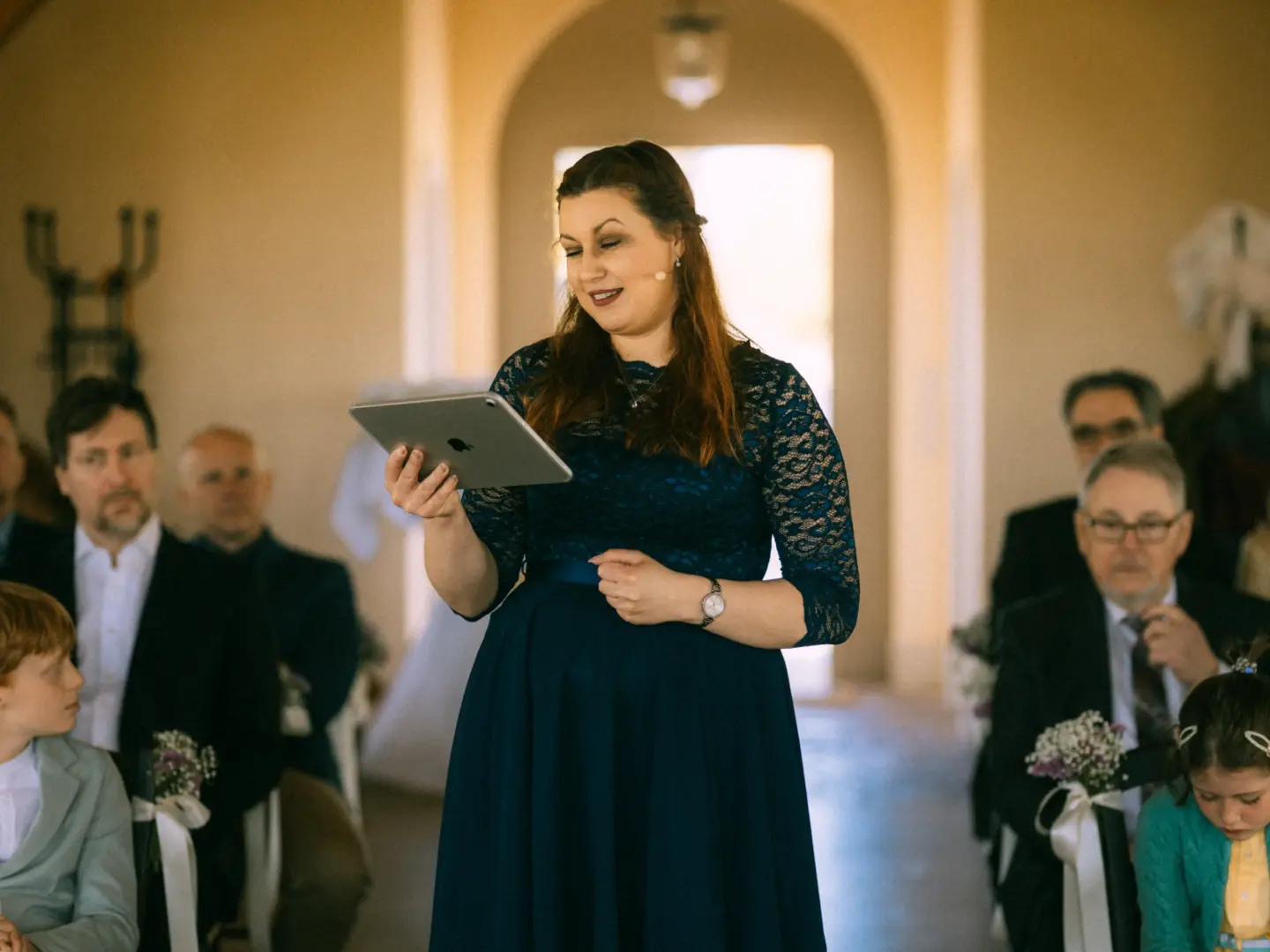 Eine Frau in einem dunkelblauen Spitzenkleid hält ein Tablet in den Händen und spricht während einer Hochzeitszeremonie zu den Gästen. Im Hintergrund sitzen aufmerksam die Hochzeitsgäste in einem hellen Raum. Authentische Aufnahme von einem Hochzeitsfotograf in Schwerin und MV.