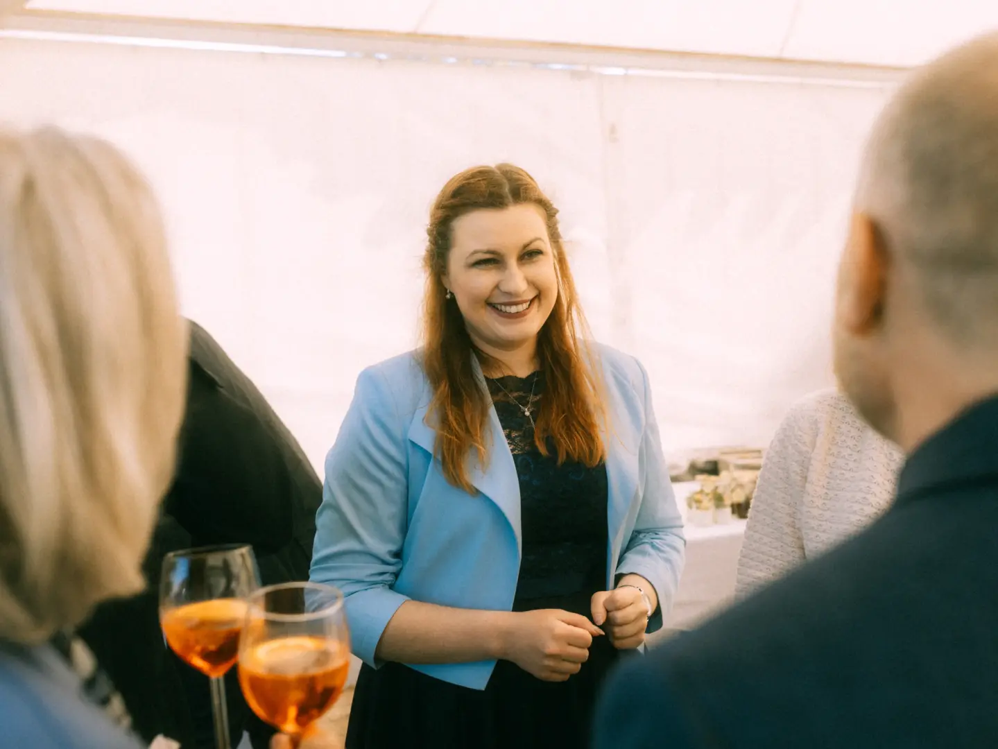 Eine lächelnde Frau in einem hellblauen Blazer und dunkelblauem Kleid unterhält sich bei einem Event mit Gästen, während sie ein Weinglas in der Hand hält. Die Atmosphäre ist herzlich und einladend, festgehalten von einem Hochzeitsfotograf in Schwerin und MV.