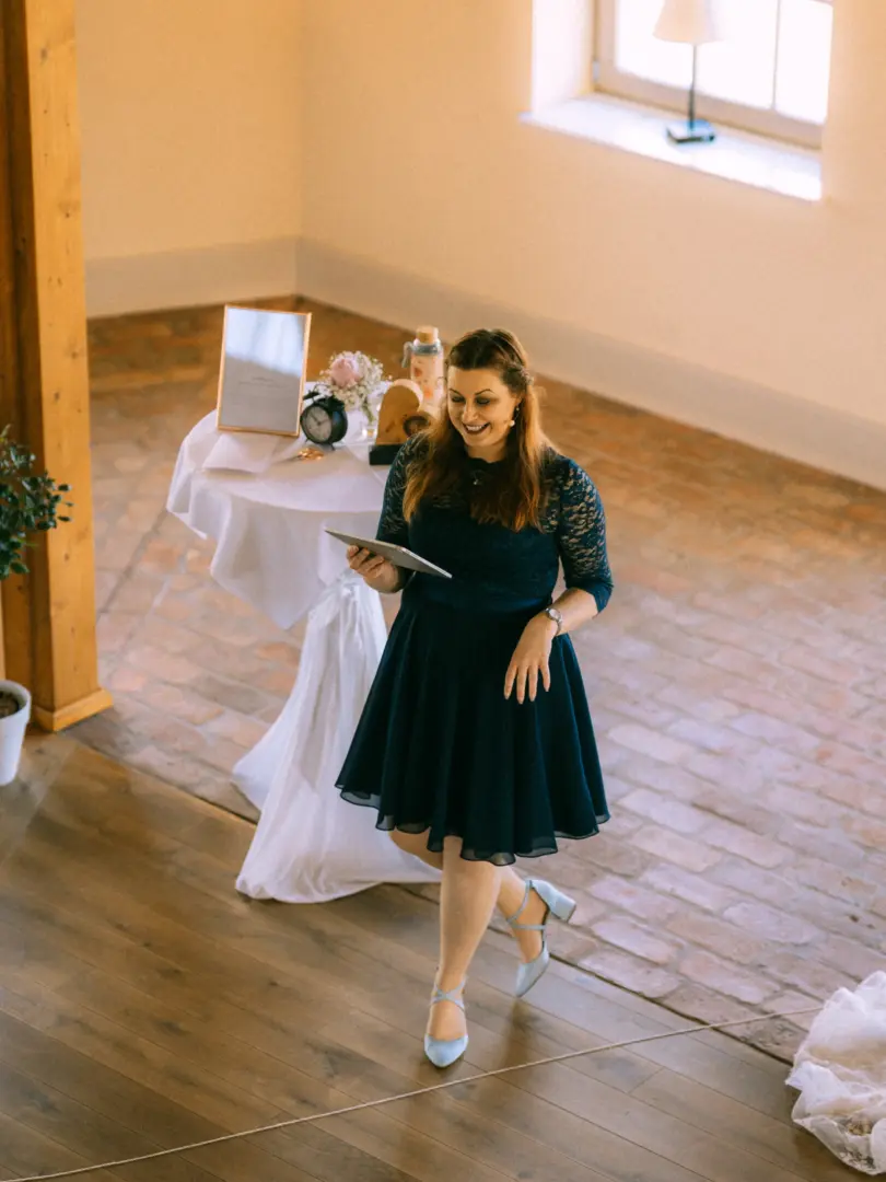 Eine lächelnde Frau in einem dunkelblauen Spitzenkleid hält ein Tablet in den Händen und präsentiert eine Hochzeitsrede. Im Hintergrund steht ein weiß gedeckter Stehtisch mit Deko. Professionelle Aufnahme von Ihrem Hochzeitsfotograf in Schwerin und MV.