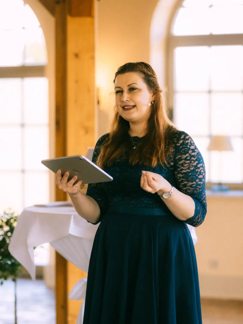 Eine Frau in einem eleganten dunkelblauen Spitzenkleid hält ein Tablet in den Händen und spricht bei einer Hochzeitsfeier. Im Hintergrund ist ein heller, festlicher Raum zu sehen. Hochzeitsfotograf in Schwerin und MV.